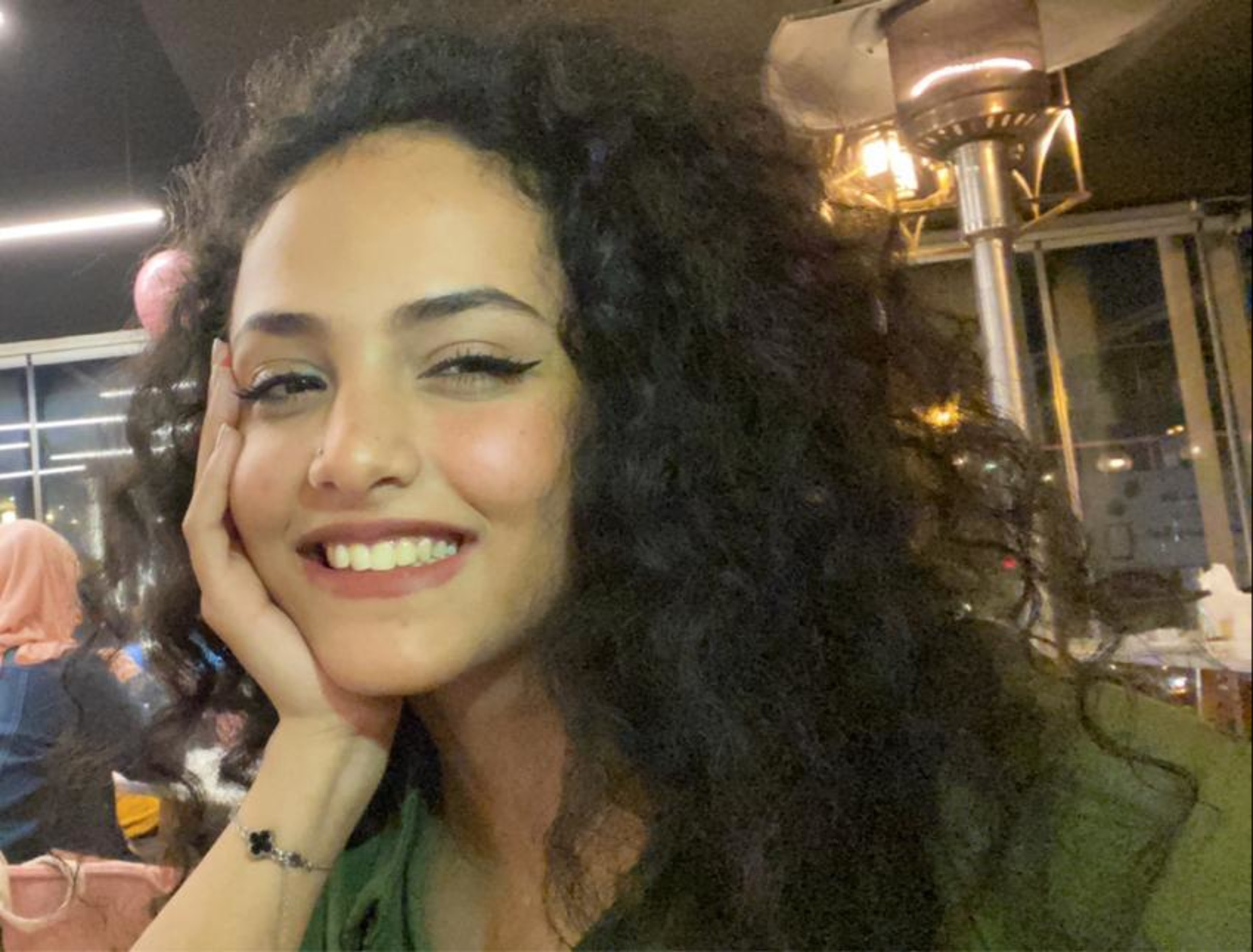 Eine junge Frau mit dunklen lockigen Haaren stützt ihr Gesicht auf ihre Hand und lacht in die Kamera.