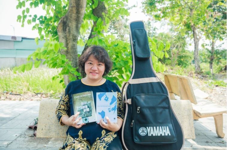 Eine lächelnde Frau mit kinnlangen dunklen Haaren sitzt auf einer Parkbank und zeigt zwei vietnamesische Bücher in die Kamera. Neben ihr ist eine Gitarrentasche.