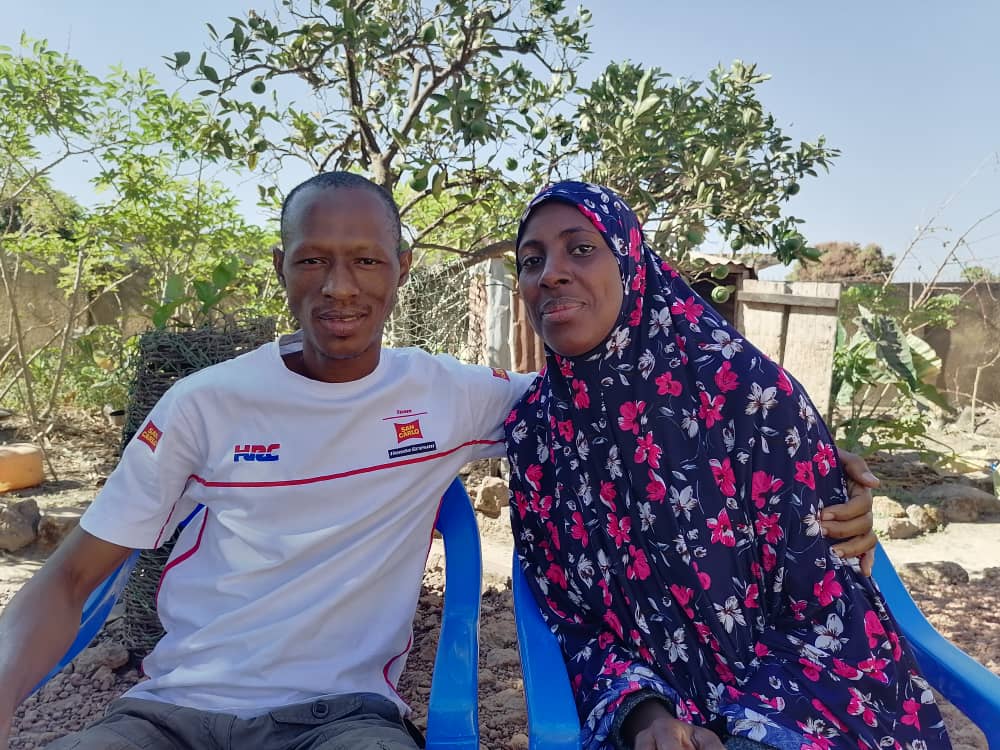 Saikou Yaya Diallo sitzt auf einem blauen Plastikstuhl. Er trägt ein weisses Sport-T-Shirt und seine Ehefrau, die neben ihm sitzt, trägt ein dunkelblaues Kleid mit pinkigen Blumen. Beide lächeln in die Kamera.
