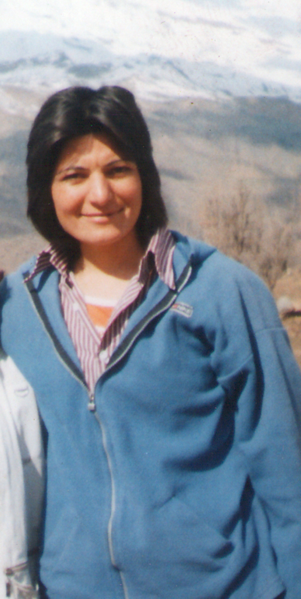 Eine Frau mit schulterlangen schwarzen Haaren und blauer Jacke lächelt in die Kamera. Die Sonne scheint und im Hintergrund befinden sich ein Tal und Berge.