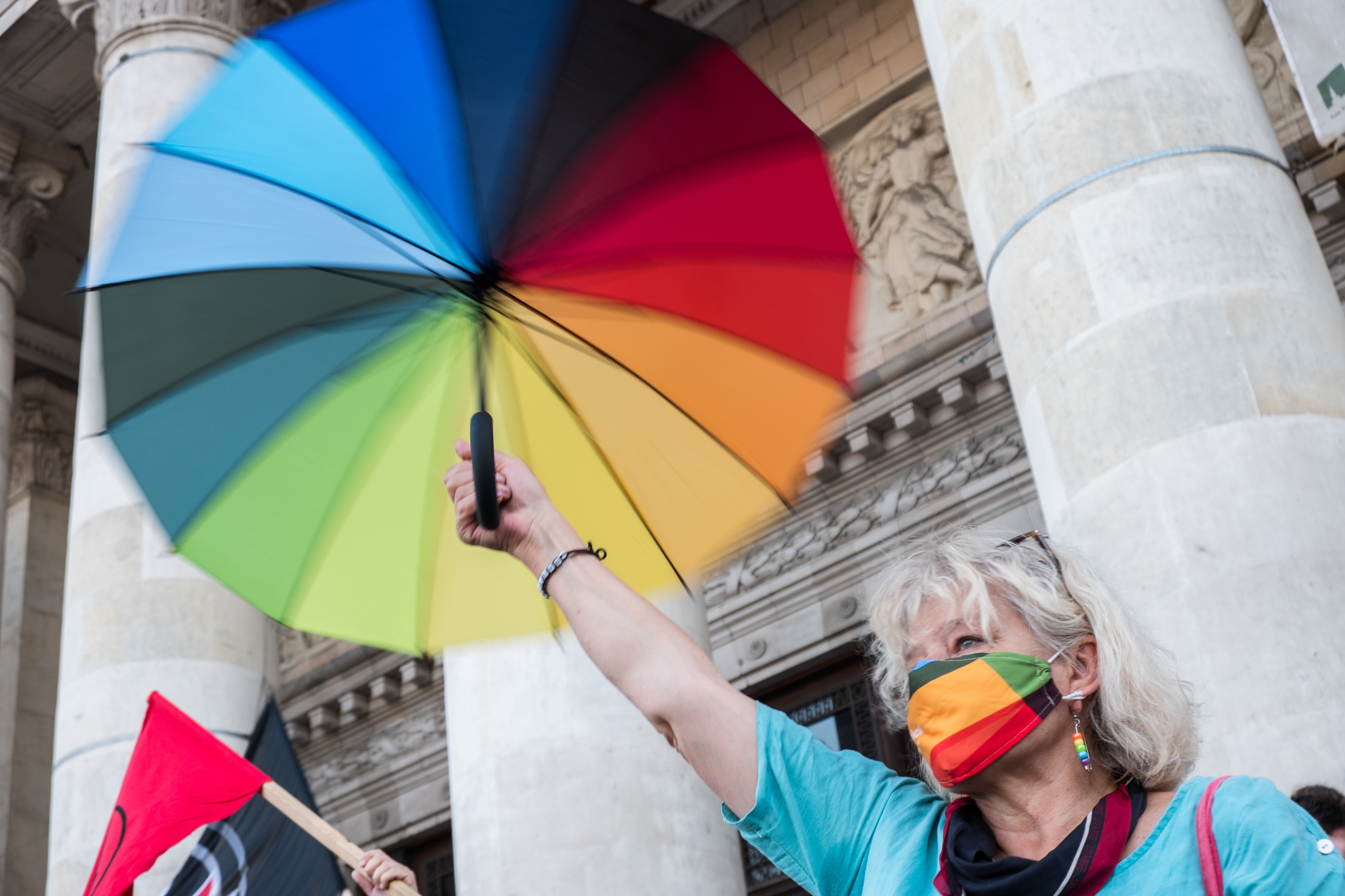Eine Frau streckt einen Regenschirm in den LGBTI-Farben in die Höhe. Sie trägt eine Gesichtsmaske und Ohrschmuck, beides auch in den LGBTI-Farben.