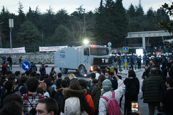Ein grauer Wasserwerfer steht im Mittelpunkt des Bildes. Im Hintergrund, vor dem Tor der Bogazici-Universität befinden sich zahlreiche Polizist_innen in Kampfmontur und im Vordergrund des Bildes hat es viele protestierende Student_innen.