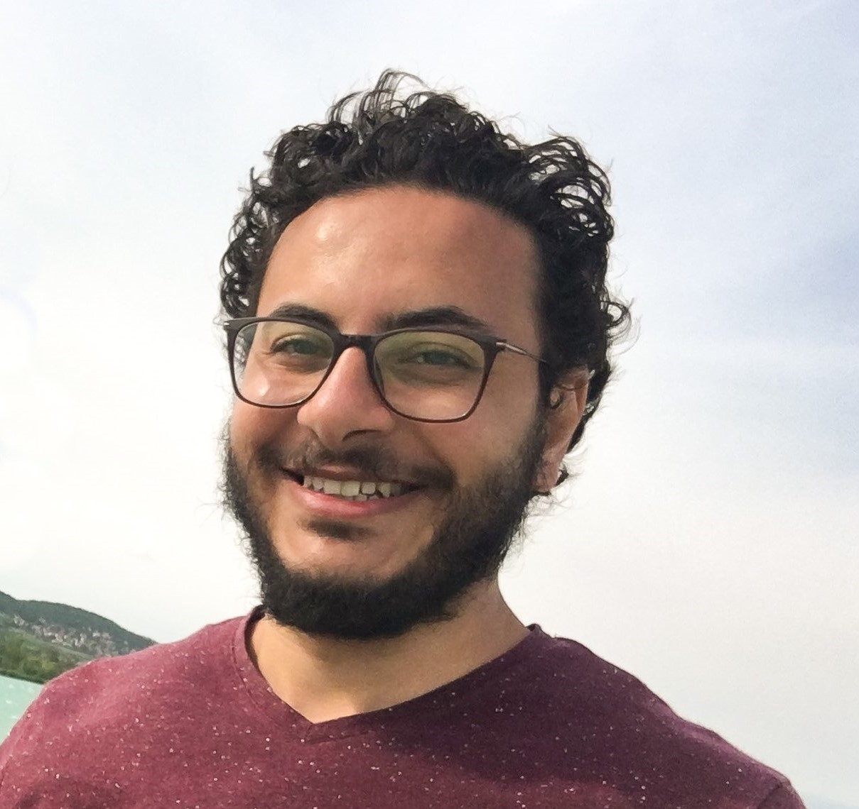 Porträt eines Mannes mit dunklen Locken und Brille, der in die Kamera lächelt.