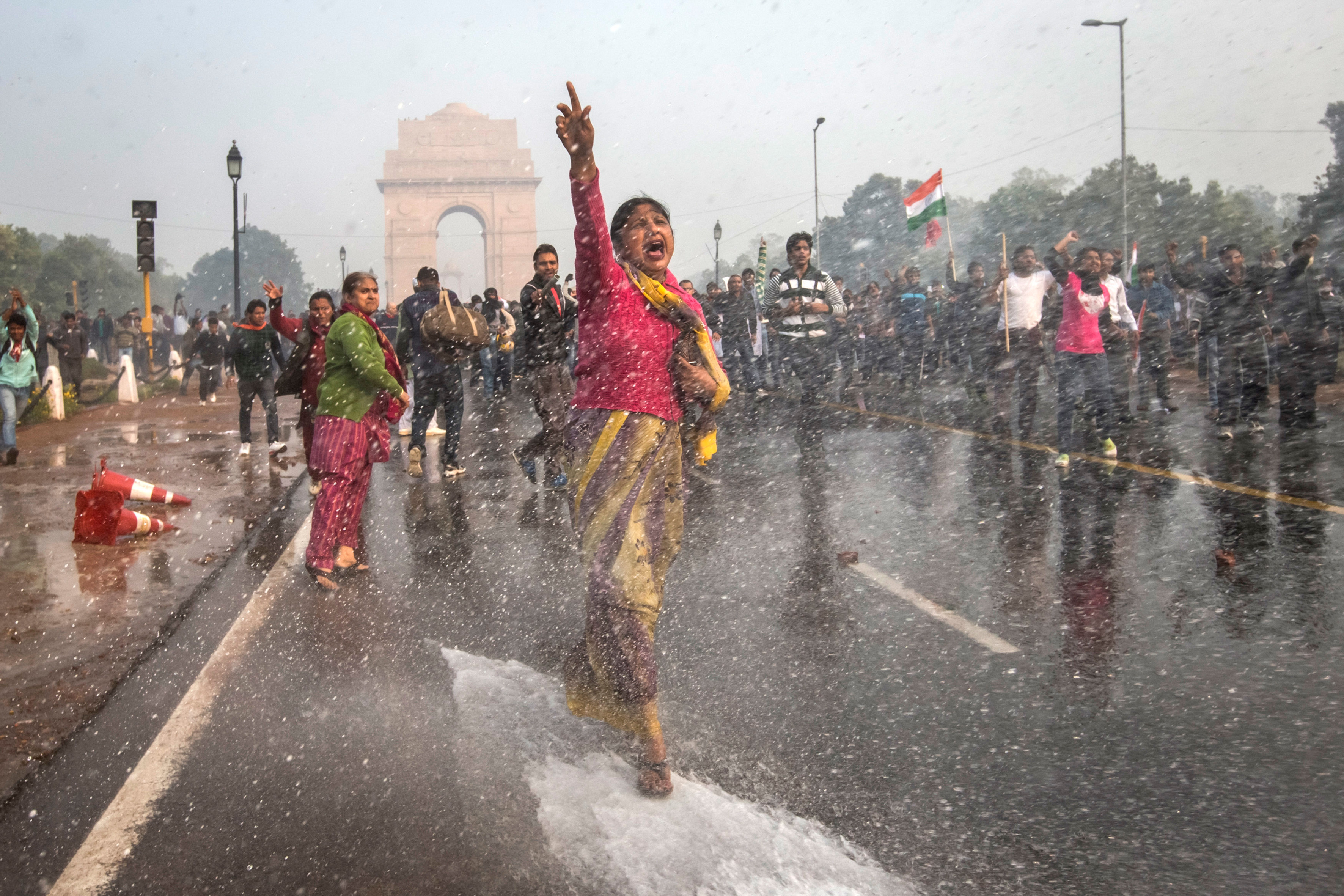 Eine Frau läuft vorm India Gate in Neu-Delhi die Straße entlang. Sie hat ihre rechte Hand erhoben und ruft etwas. Dabei treffen sie Wassertropfen, potenziell von einem Wasserwerfer. Hinter ihr befinden sich weitere Protestierende.