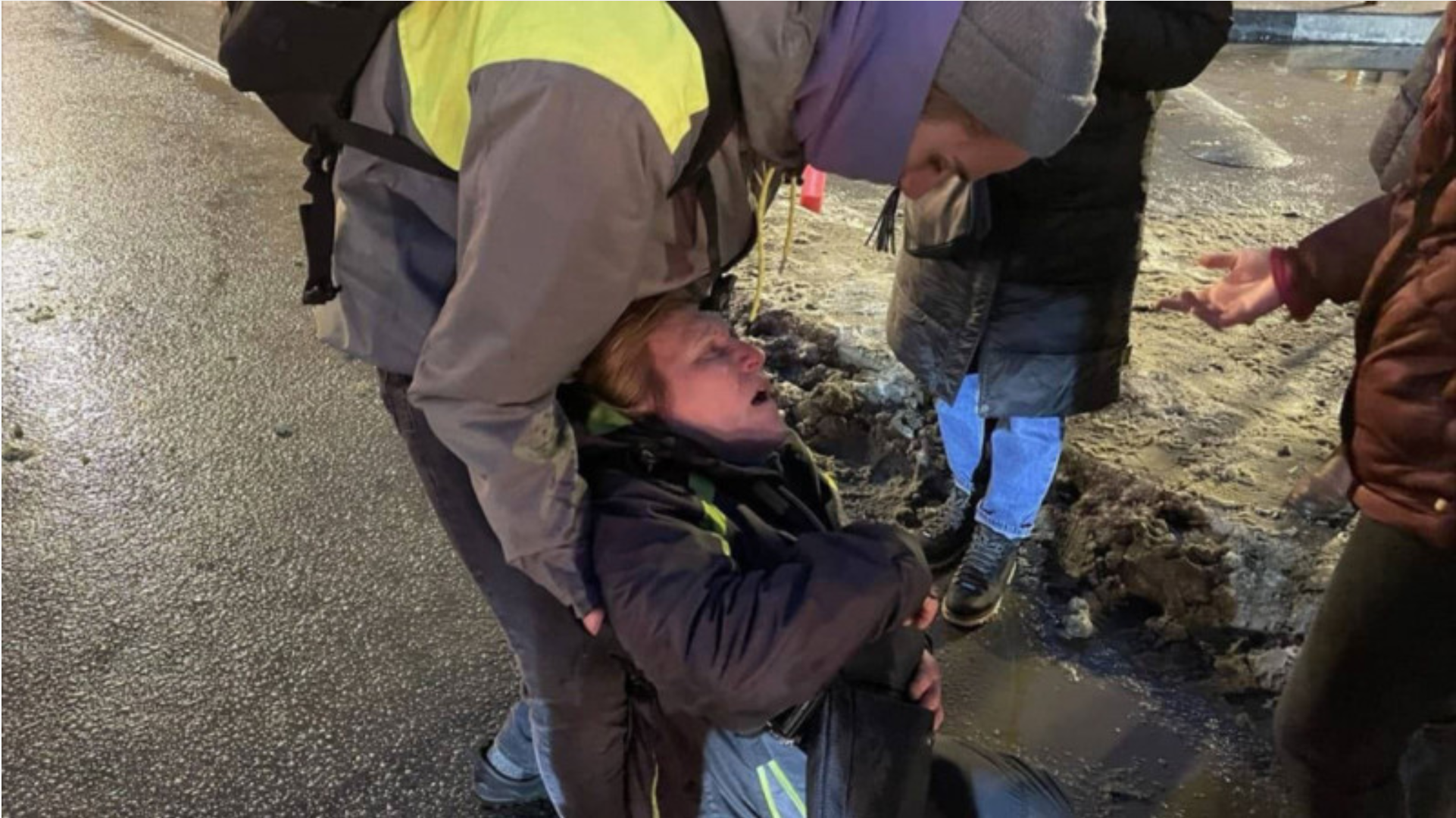 Frau beugt sich über über die am Boden liegende Margarita Yudina und stützt ihren Rücken. Das Gesicht von Margarita Yudina ist schmerzverzerrt.