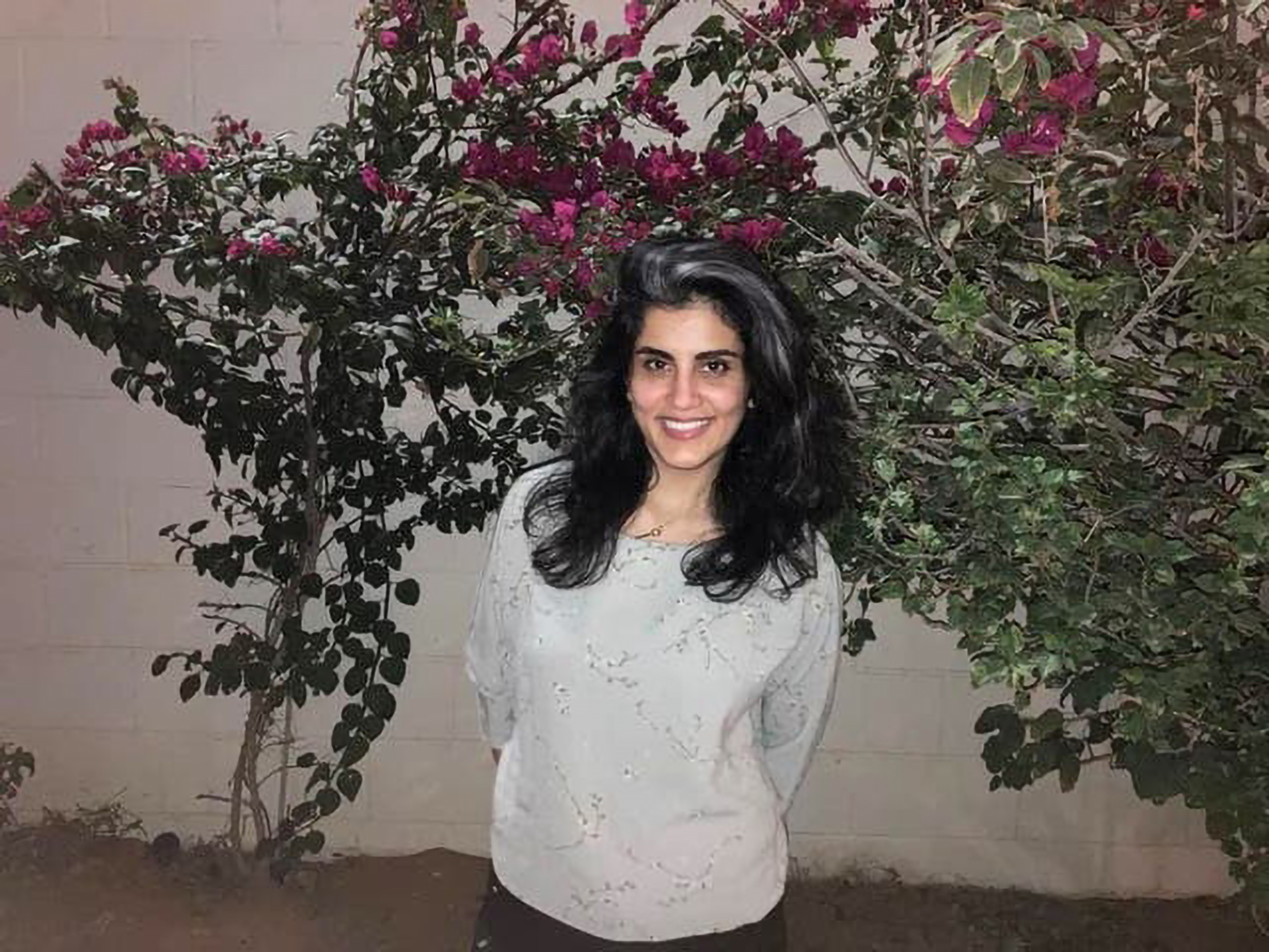 Loujain Al-Hathloul lächelt in die Kamera, sie trägt ihr teilweise leicht ergrautes Haar offen. Sie steht vor einer Mauer, die mit blühenden Blumensträuchern bedeckt ist.