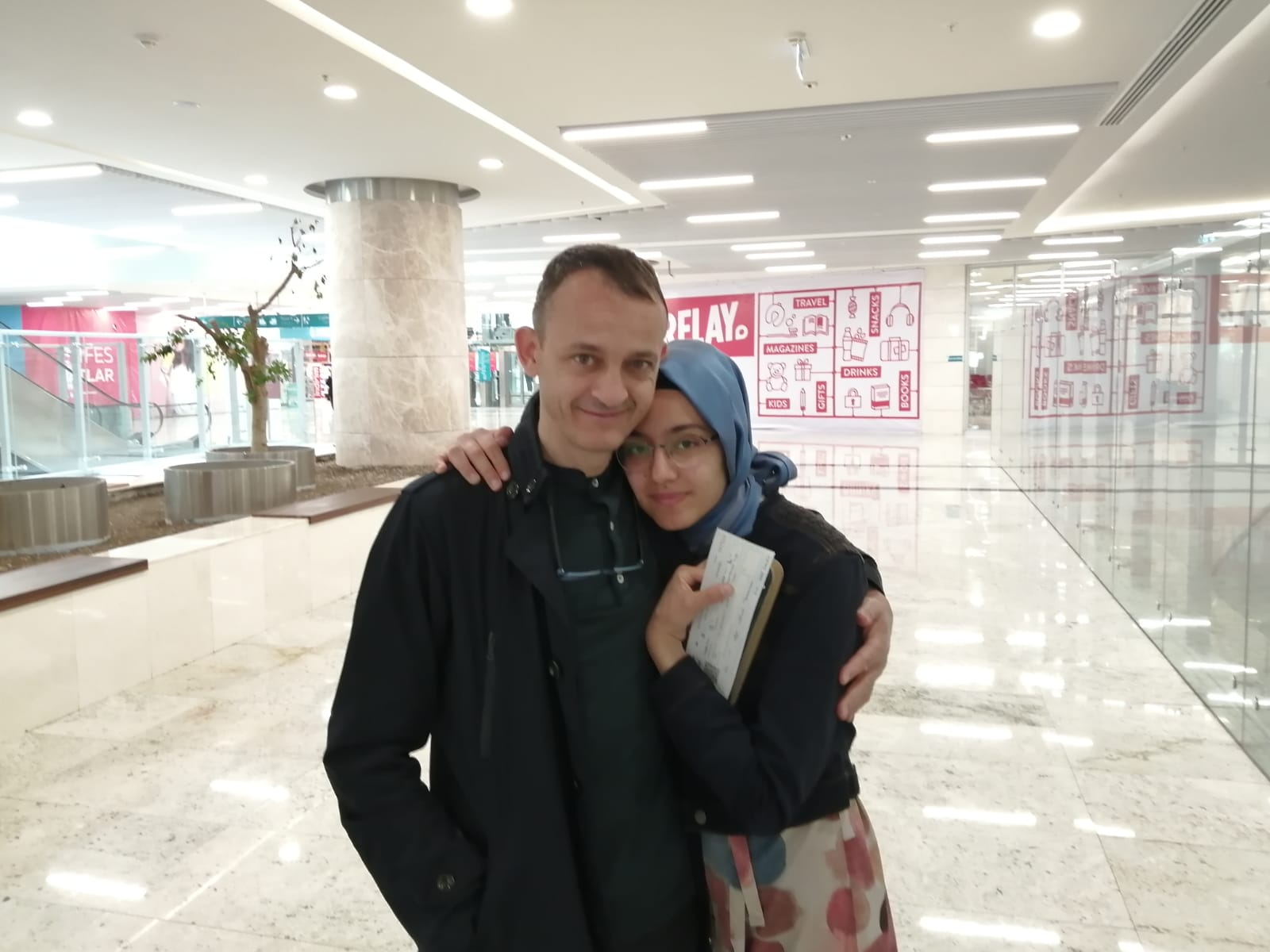Hüseyin Galip Küçüközyiğit und seine rechts neben ihm stehende Tochter umarmen sich und blicken in die Kamera. Sie stehen allein in einem breiten, hell erleuchteten Flur in einem Einkaufszentrum oder einem Flughafen.