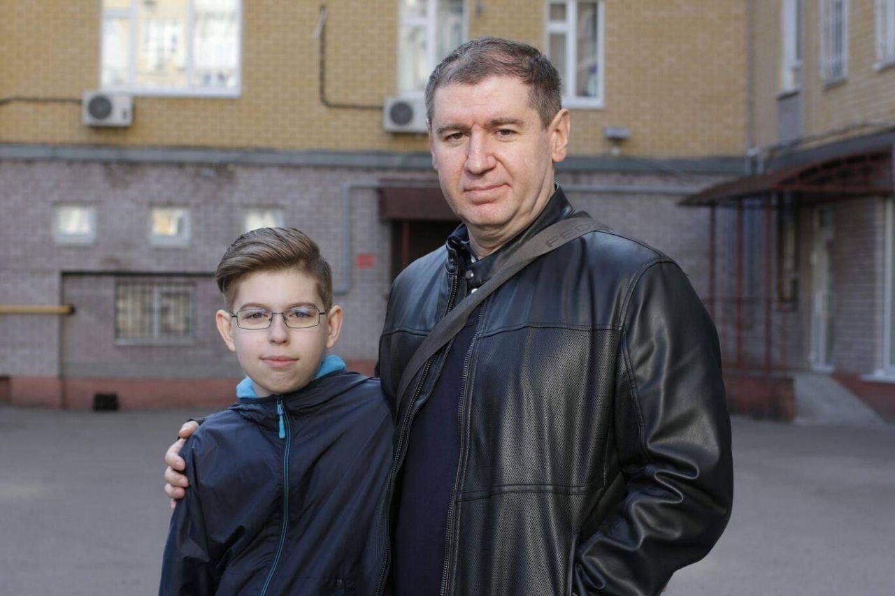Ein älterer Mann und ein Kind posieren für die Kamera. Sie halten sich umarmt. Im Hintergrund ist eine Hausfassade zu erkennen.