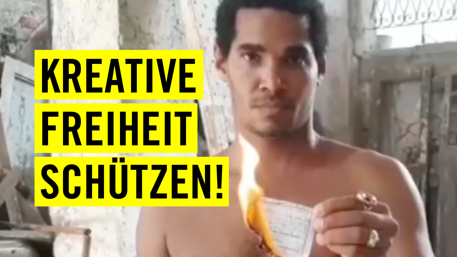 Film-Still von einem Mann, der Richtung Kamera schaut und einen Zettel, der in Flammen steht in der Hand hält. Links im Bild ist der Schriftzug "Kreative Freiheit schützen!" auf drei gelben Balken.
