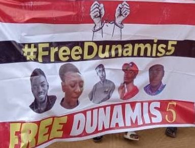 Zu sehen ist ein Banner mit der Aufschrift "Free Dunamis" und den Portraits der fünf Aktivisten, das offensichtlich von mehreren Personen getragen wird (von denen nur Hände und Füße zu sehen sind).