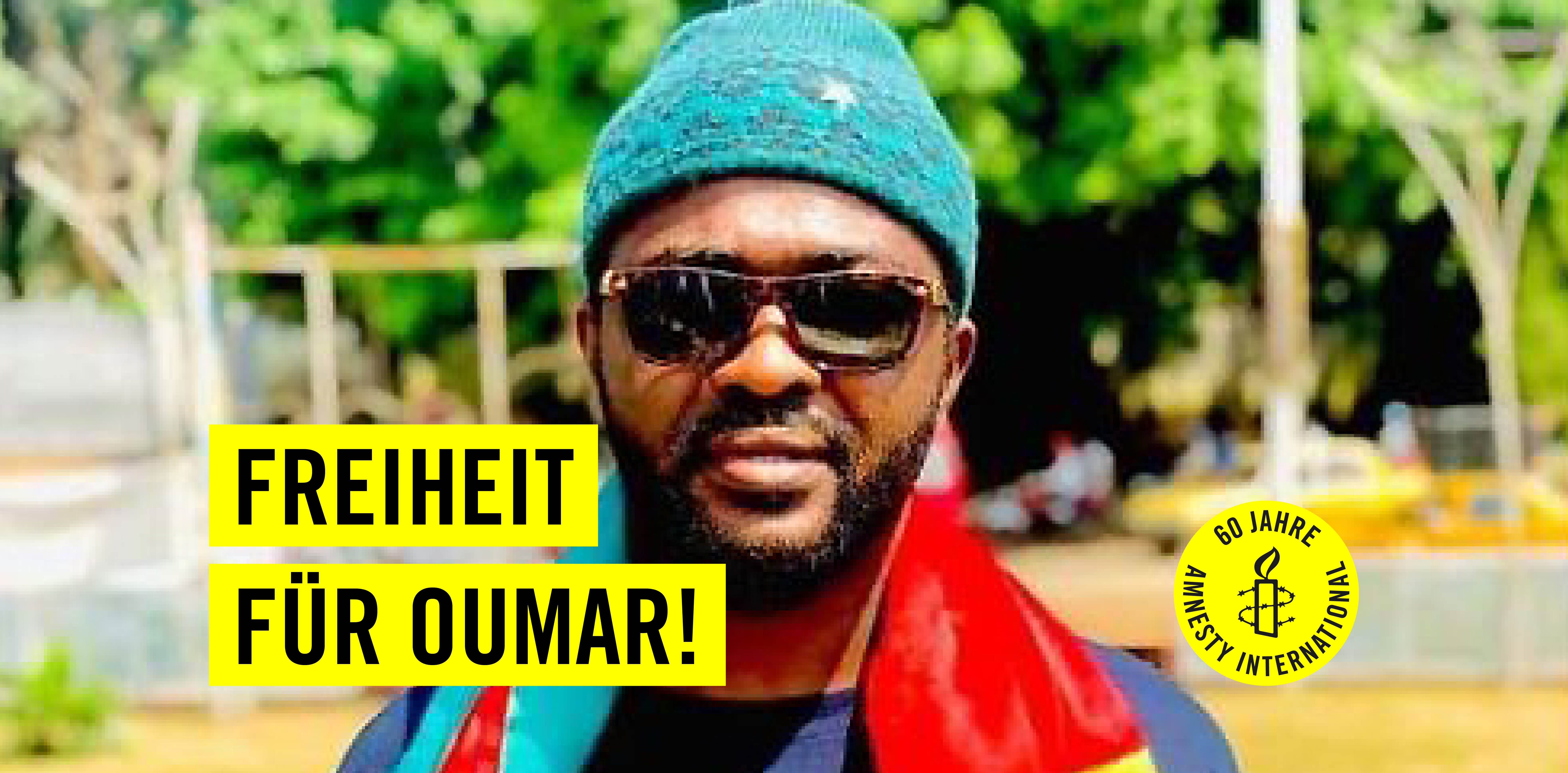 Ein Mann mit Sonnenbrille und Mütze. Auf gelben Balken steht in schwarzer Schrift: Freiheit für Oumar! Auf einem gelben Kreis ist eine Kerze, die von Stachesdraht umschlungen wird, zu sehen und es steht "60 Jahre Amnesty International" geschrieben. 