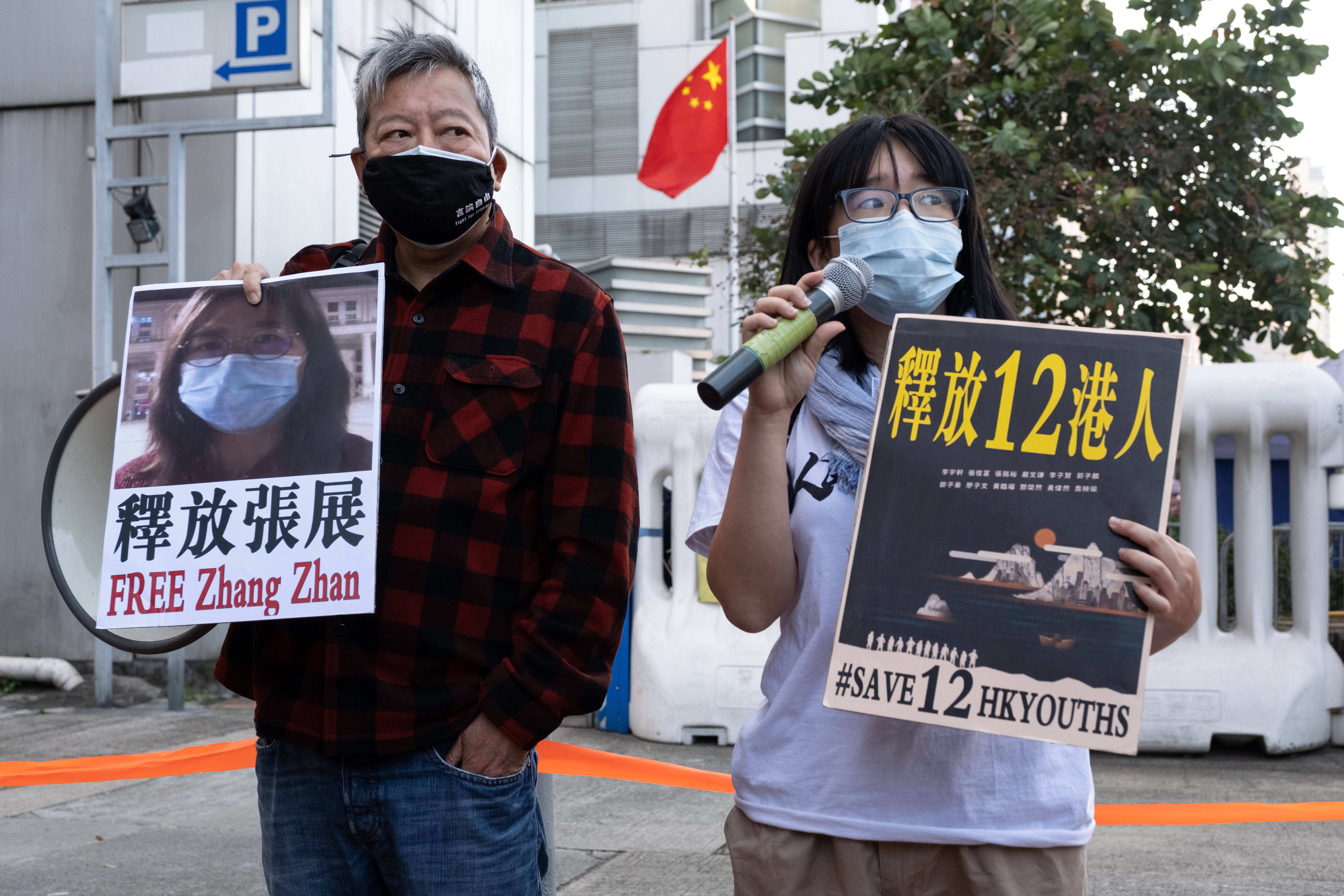 Das Bild zeigt zwei Menschen, die Protestschilder hochalten, sie stehen vor einem Gebäude mit chinesischer Flagge, auf einem Plakat ist das Foto einer Frau zu sehen sowie der Schriftzug Zhang Zhan