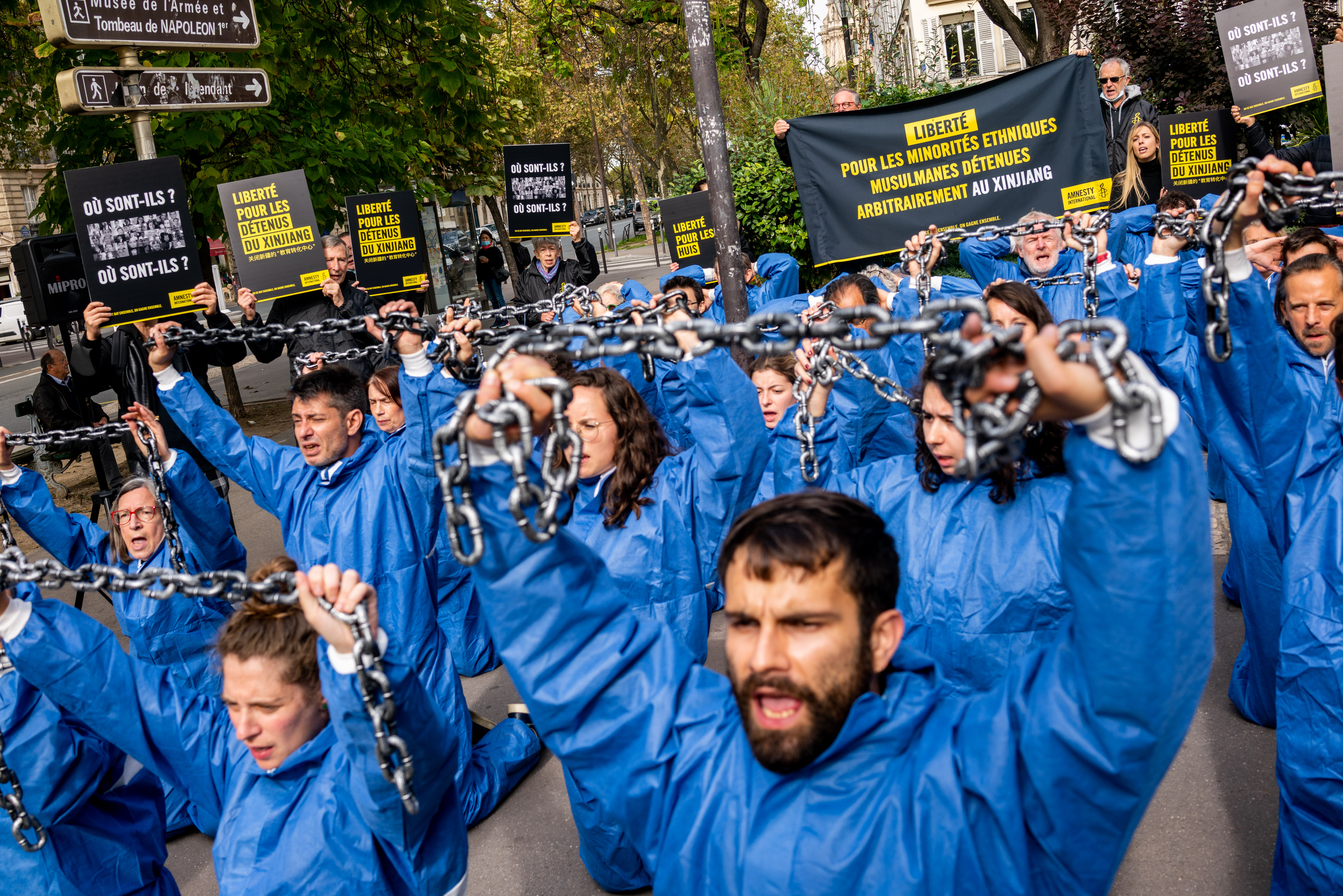 Das Bild zeigt eine Protestaktion, mehrere Menschen mit blauen Overalls knien auf dem Boden und halten schwere Eisenketten in de Höhe