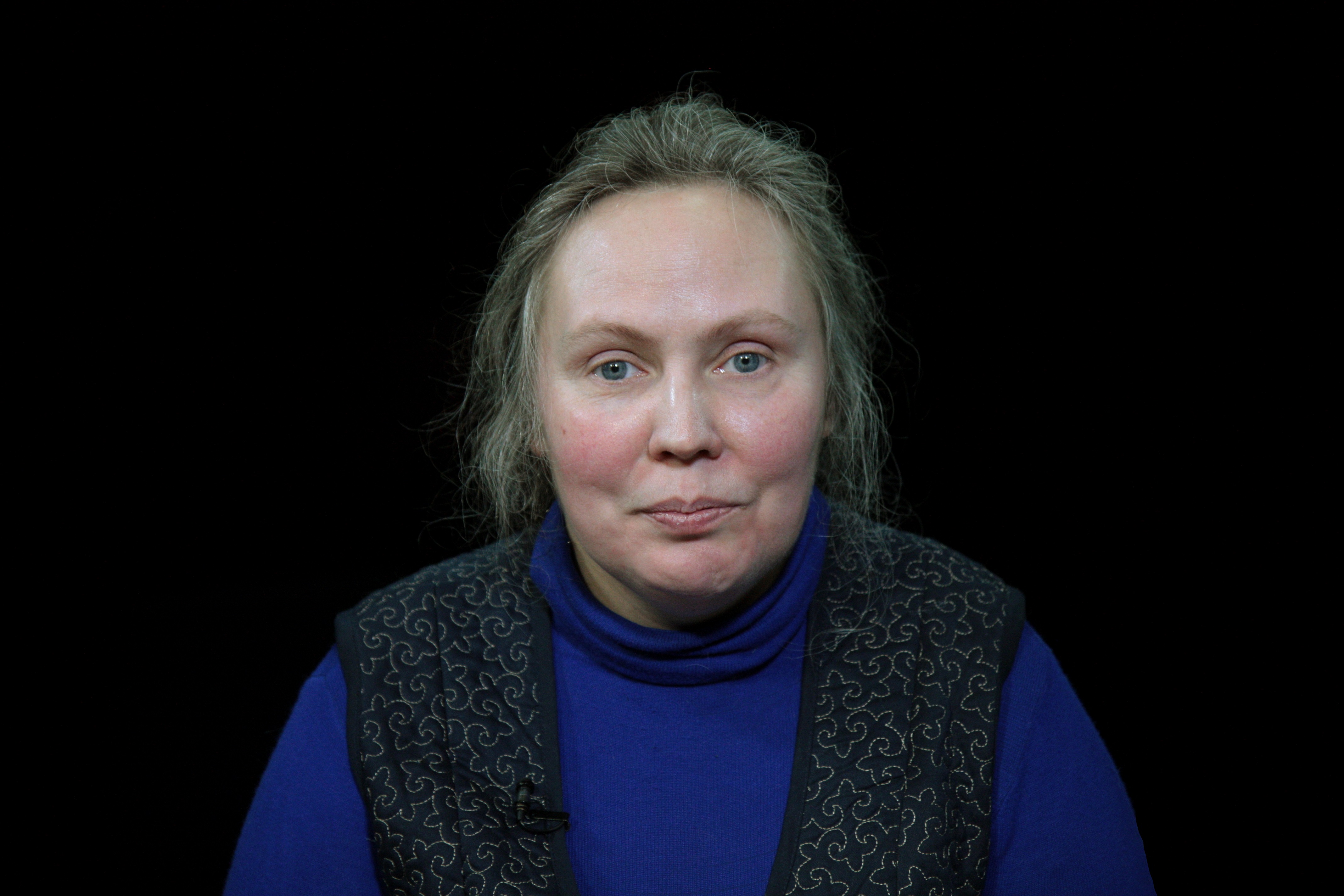Portraitfoto einer Frau auf schwarzem Grund. Die Frau hat helle, zusammengenommene Haare, blaue Augen und sie trägt einen dunkelblauen Rollkragenpullover.