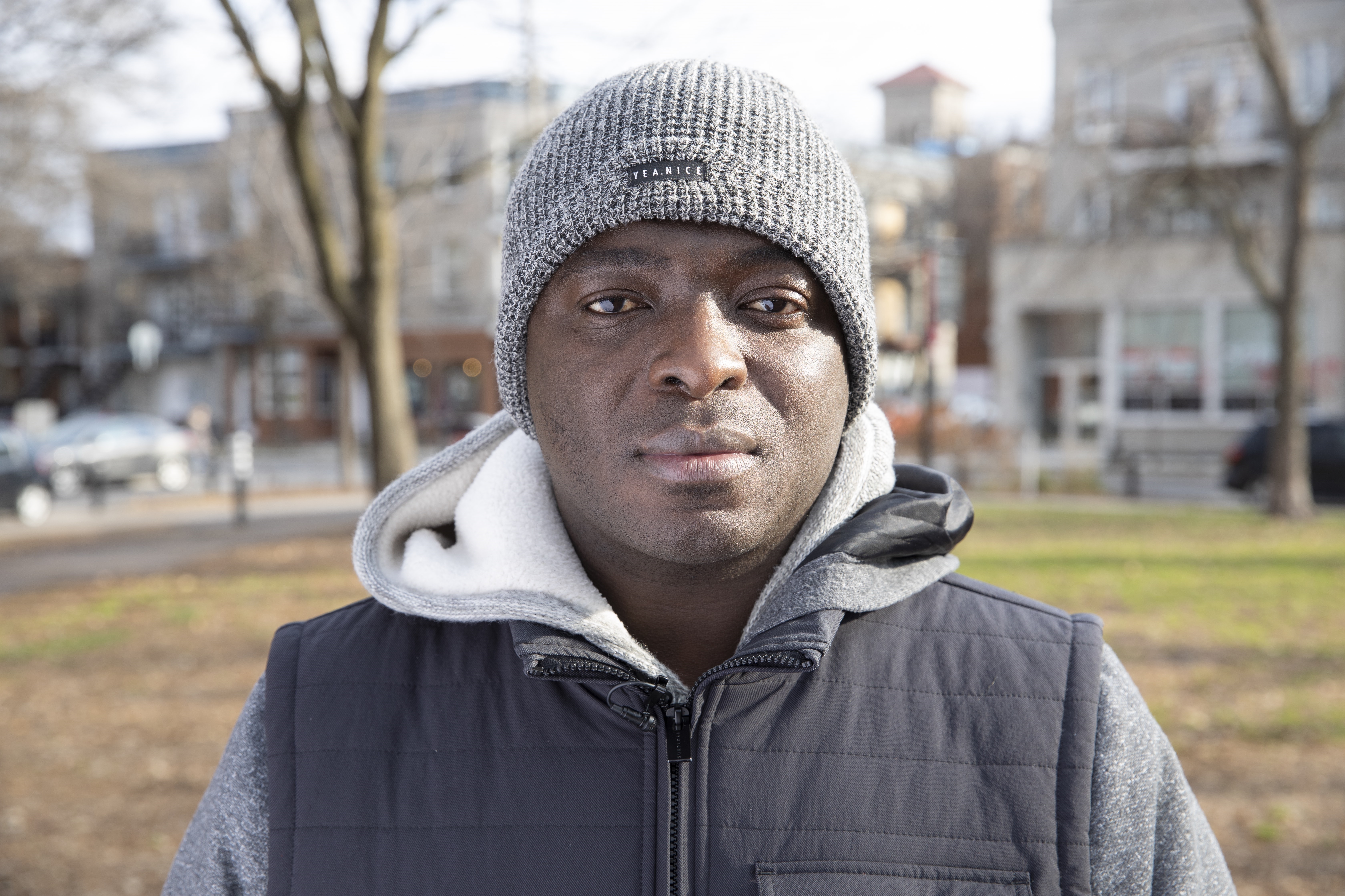 Portraitfoto eines Mannes draussen, er trägt eine graue Mütze und trägt einen grauen Hoodie mit einer schwarzen Veste darüber. Er schaut ernst in die Kamera.