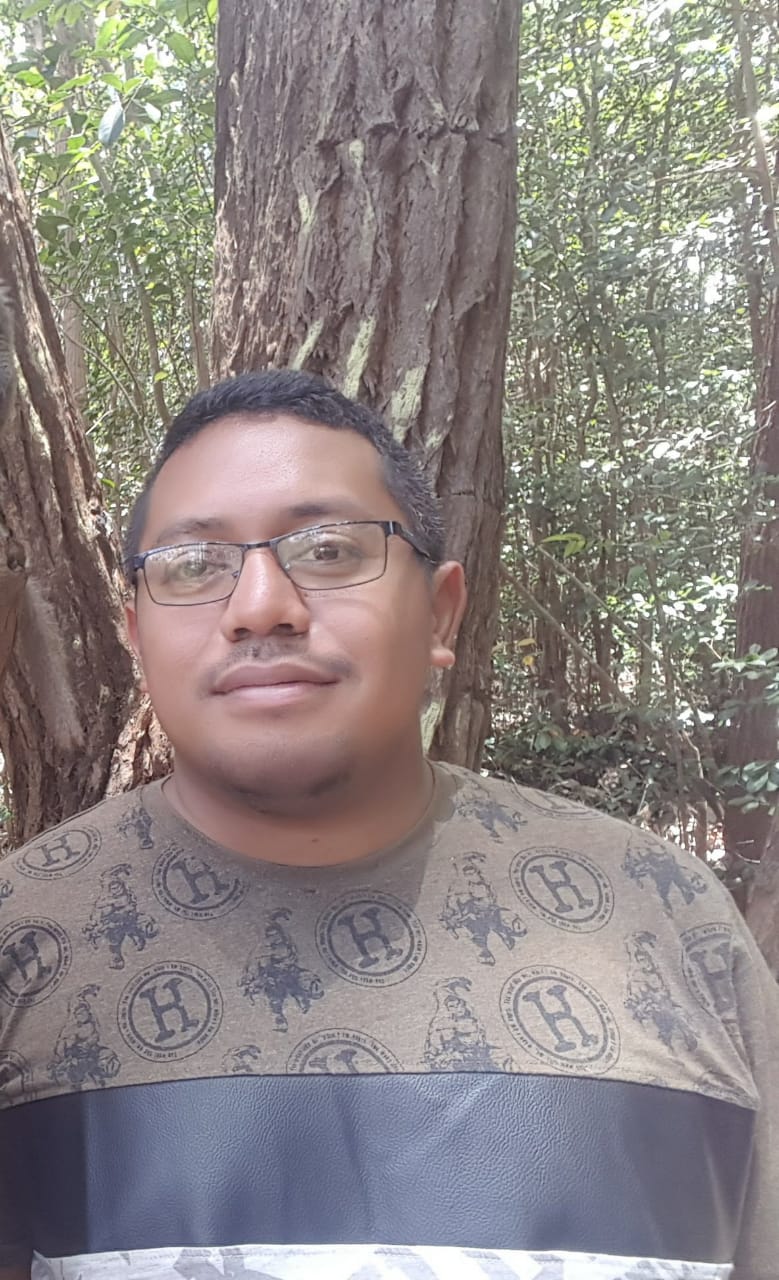 Portraitfoto eines Mannes, der eine Brille trägt und kurze schwarze Haare hat. Im Hintergrund sind tropische Bäume und Blätter.