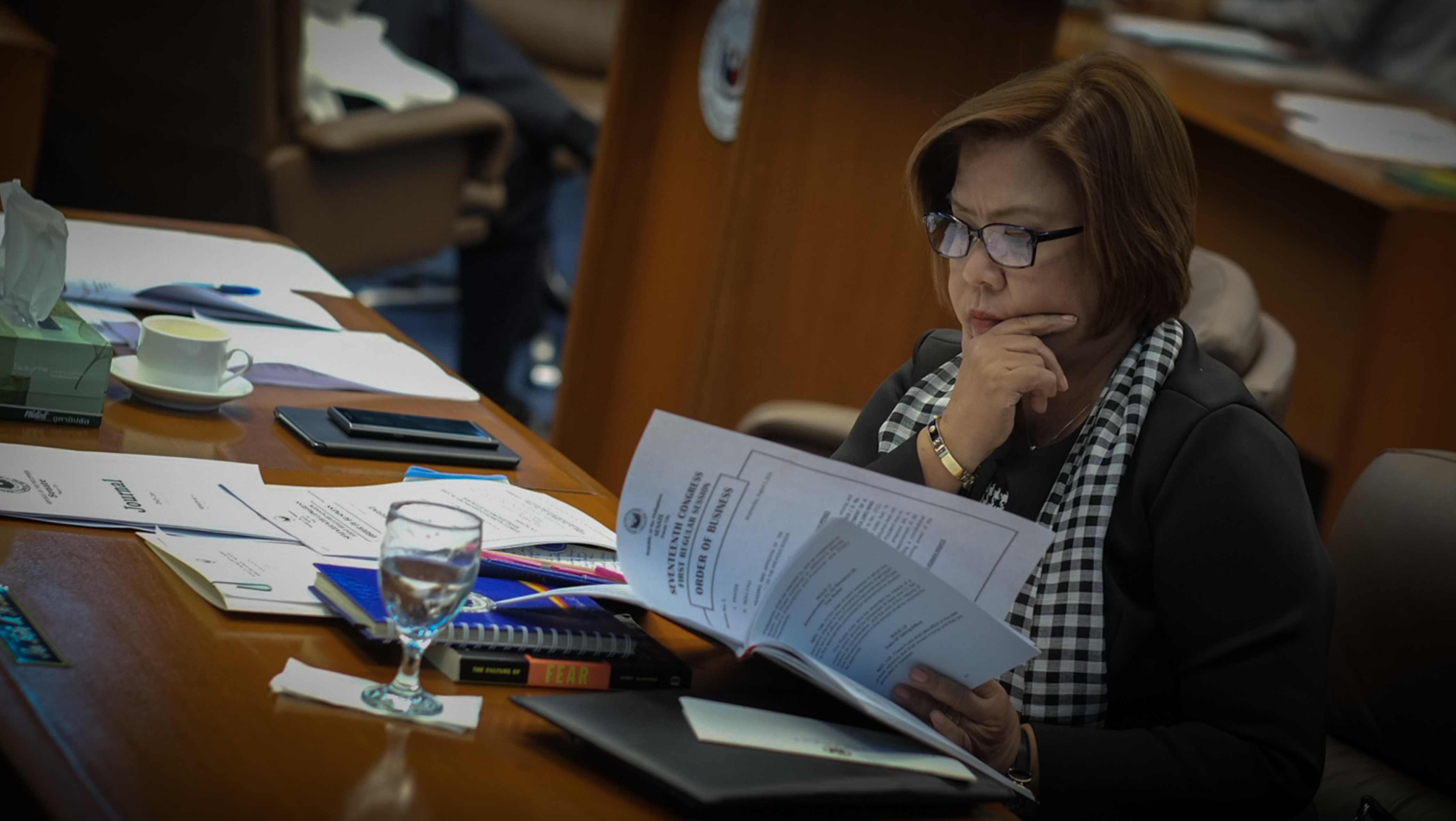 Foto einer Frau mit Brille, die Blätter in der Hand hält und am lesen ist. Die andere Hand legt sie an ihr Kinn.