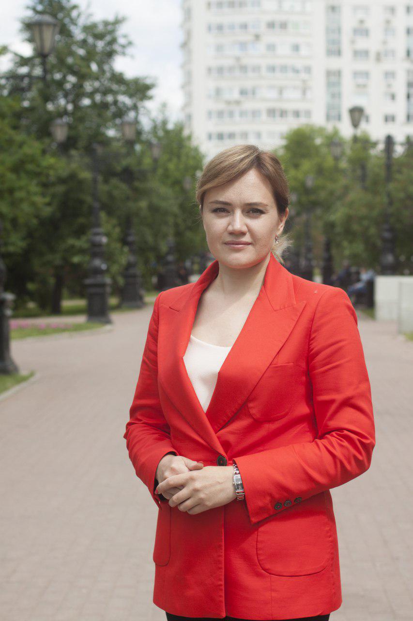 Portraitfoto einer blonden Frau. Sie trägt einen roten Blazer und ist streng frisiert und geschminkt. Sie schaut ernst in die Kamera. Im Hintergrund ist eine Stadt mit einem Hochhaus und grünen Bäumen.