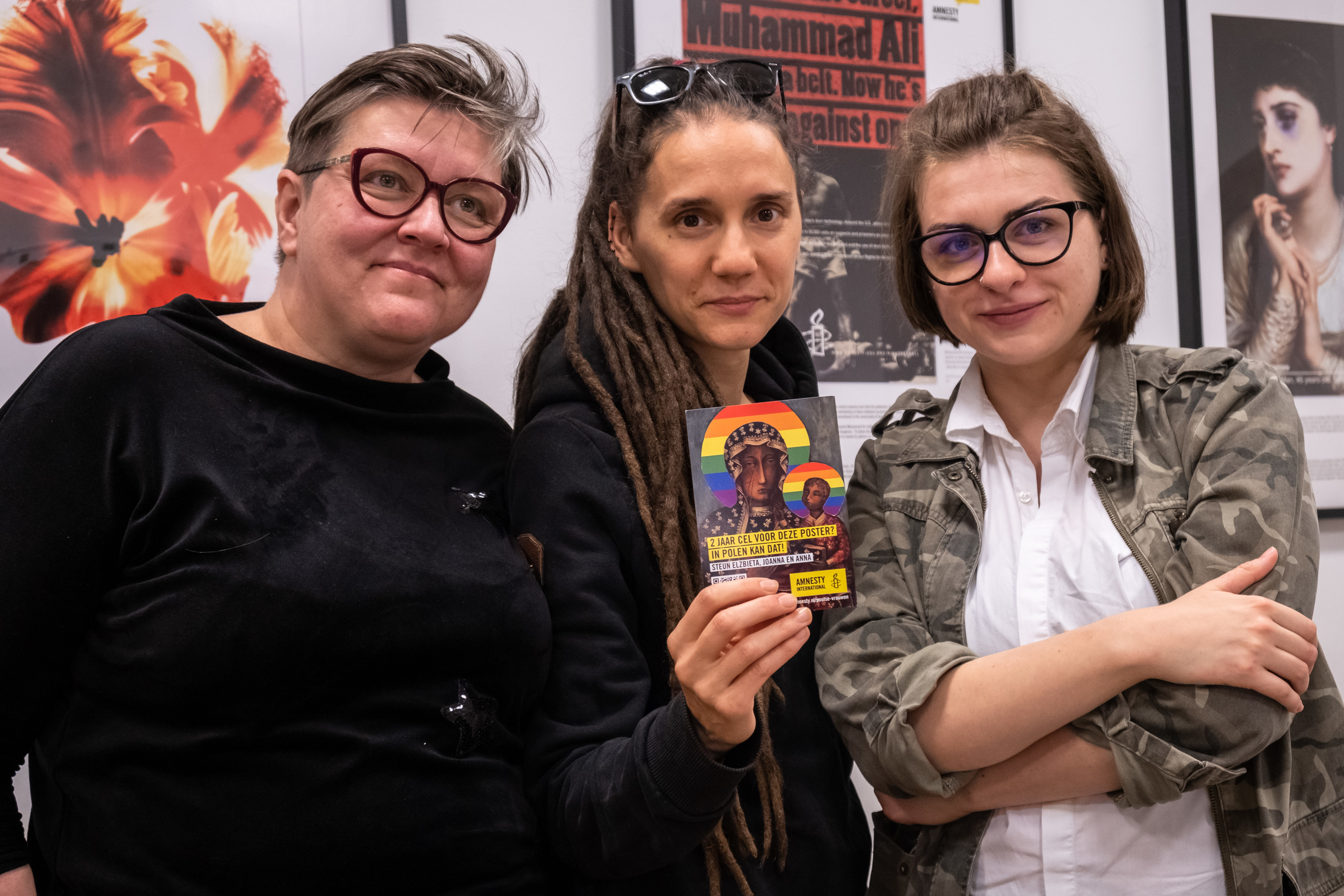 Foto von drei Frauen, die mittlere hält eine Postkarte mit dem Motiv der Jungfrau Maria in der Hand, deren Heiligenschein regenbogenfarbig ist