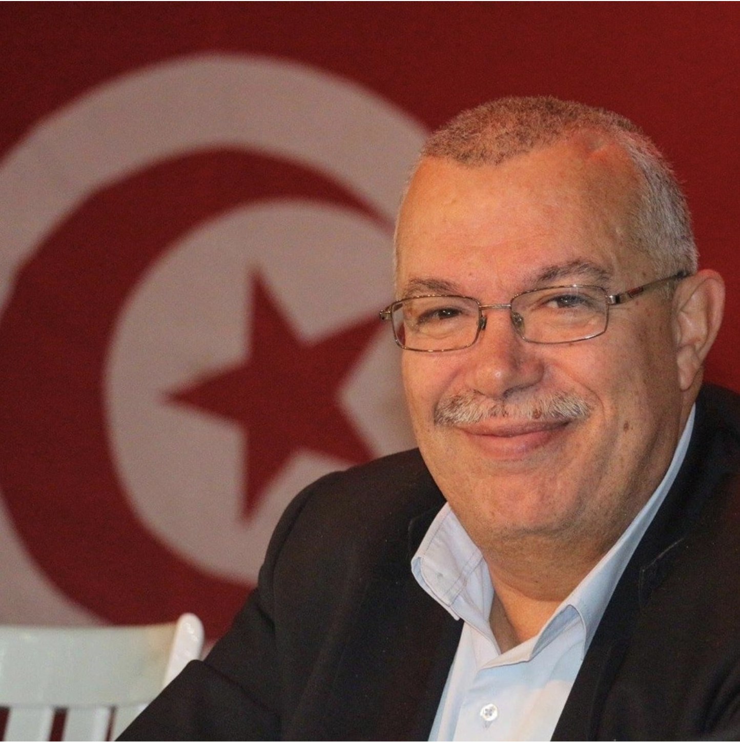 Porträtfoto von Noureddine Bhiri, der eine Brille trägt und in die Kamera lächelt. Im Hintergrund hängt die tunesische Flagge an der Wand.