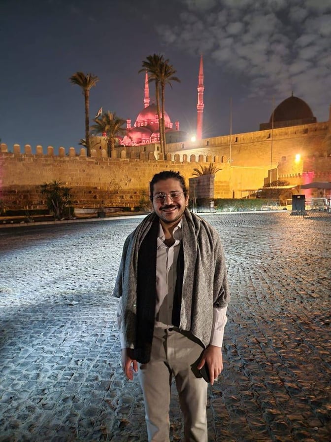 Patrick George Zaki steht nachts auf einem Platz, im Hintergrund steht eine hell erleuchtete Moschee.