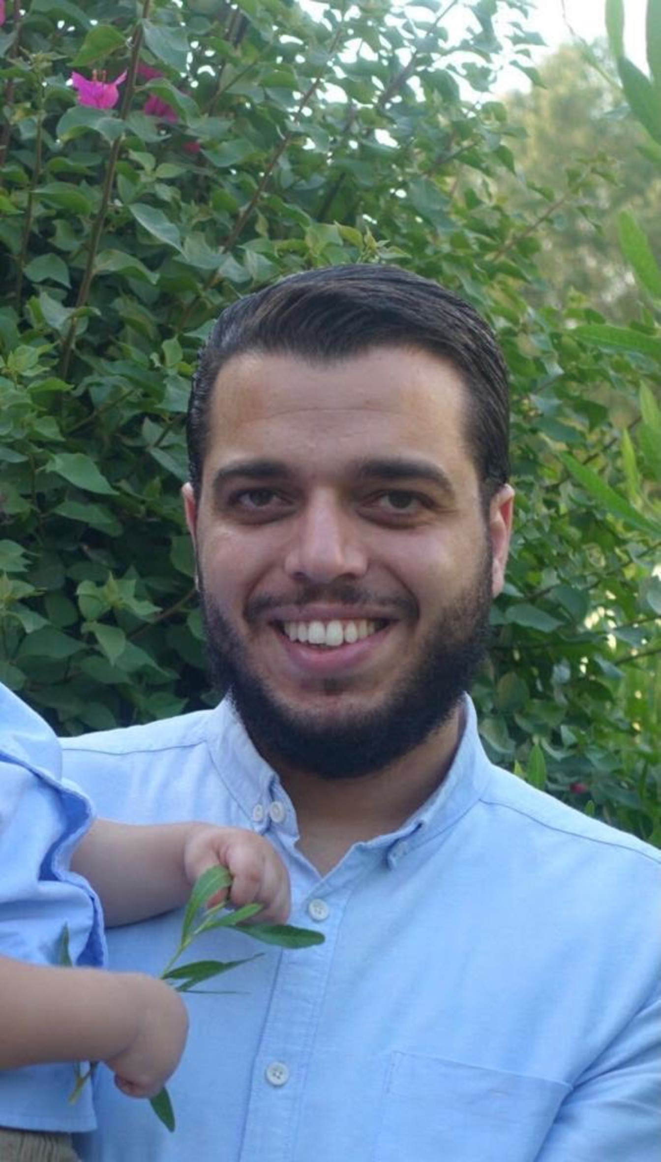 Das Foto zeigt Abdullah Ibhais mit Vollbart vor Büschen stehend in einem Garten. Er trägt ein Hemd und lächelt in die Kamera und hält auf seinem rechten Arm ein kleines Kind.