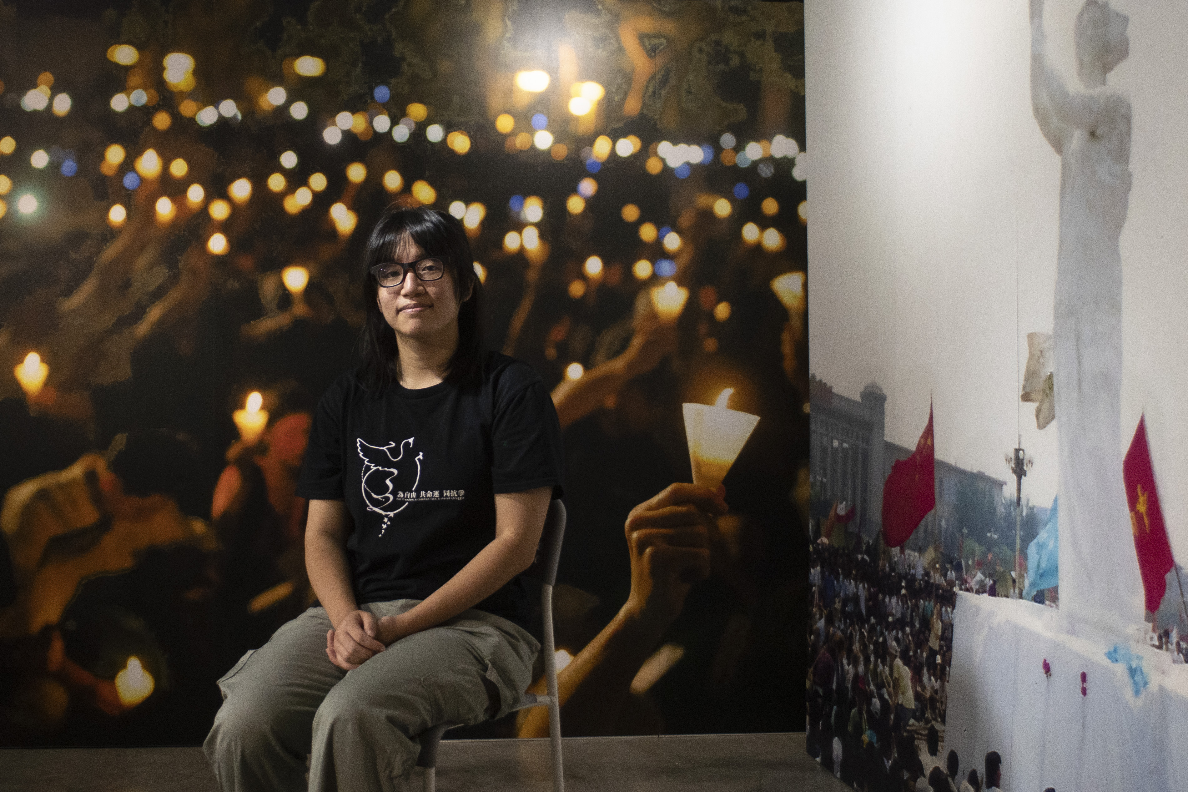 Das Bild zeigt eine junge Frau, die auf einem Stuhl sitzt und in die Kamera schaut, im Hintergrund ist ein Poster auf dem Lichter zu sehen sind. 