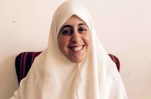 Porträtfoto von Aisha el-Shate, die auf einem Stuhl sitzt und ein Kopftuch trägt und in die Kamera lächelt.