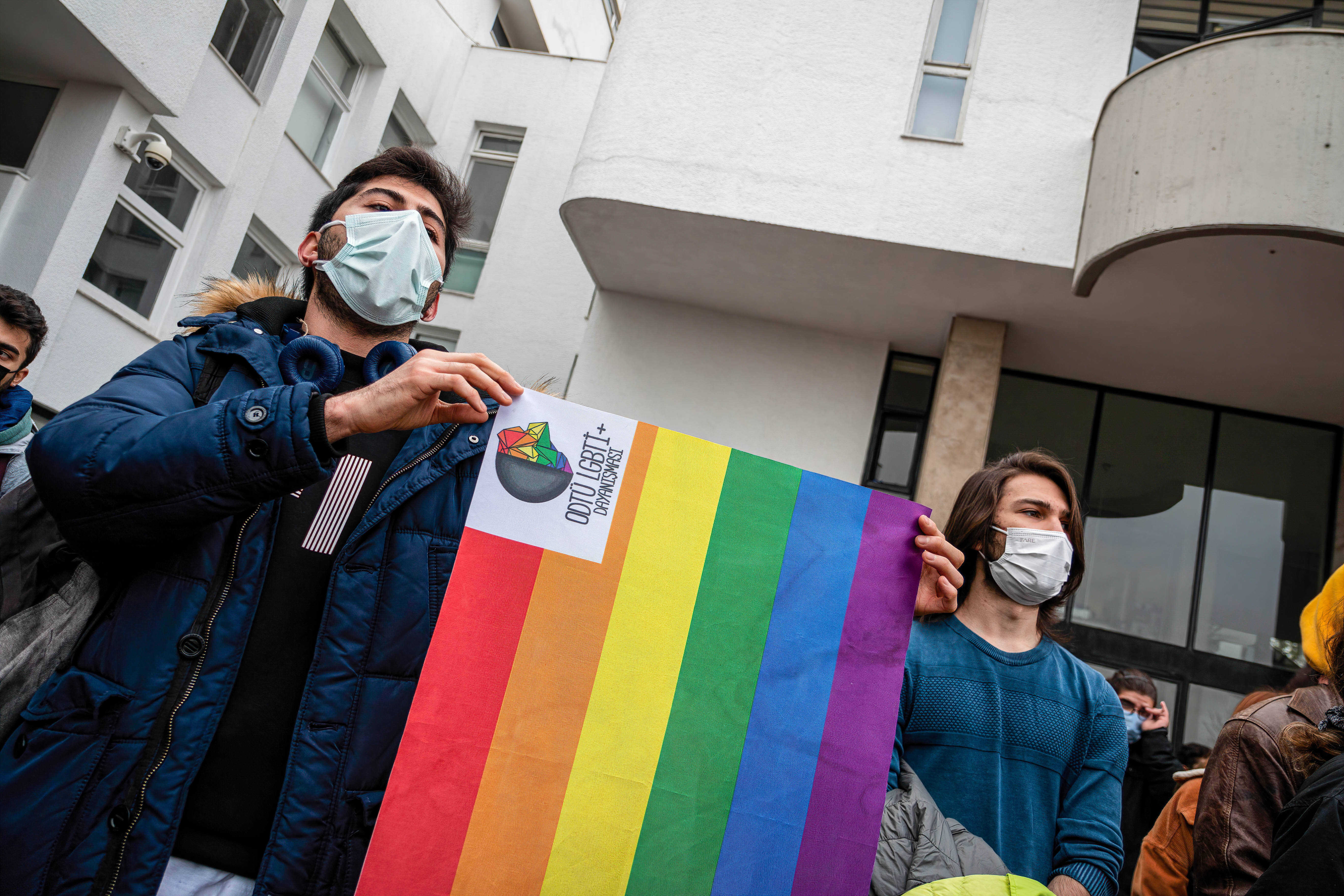 Zwei Männer mit Virenschutzmaske halten vor einem Gebäude eine Regenbogen-Fahne hoch.