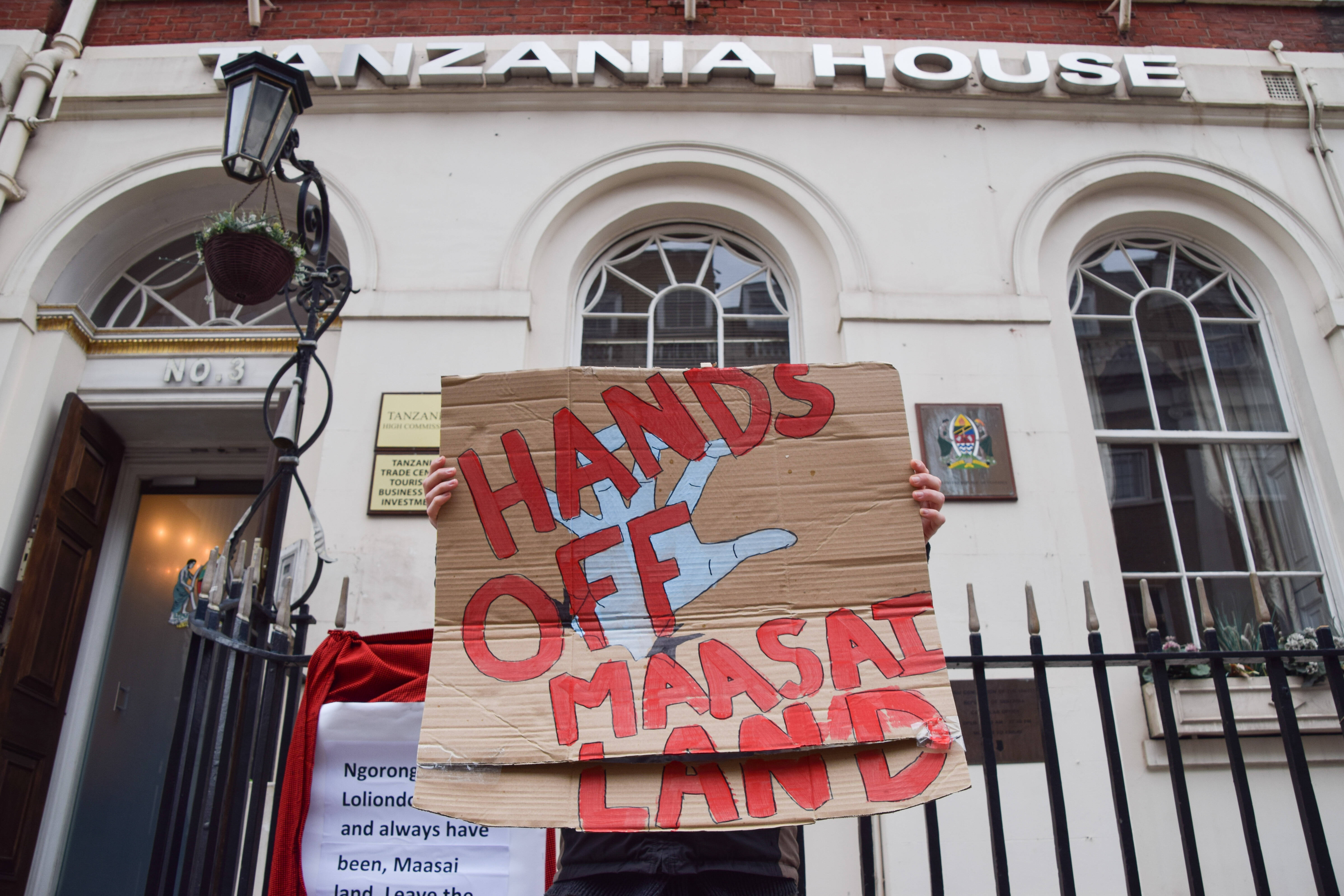 Das Bild zeigt eine Person, die vor einem Gebäude steht und ein Schild in der Hand hält: "Hands off Maasai Land"