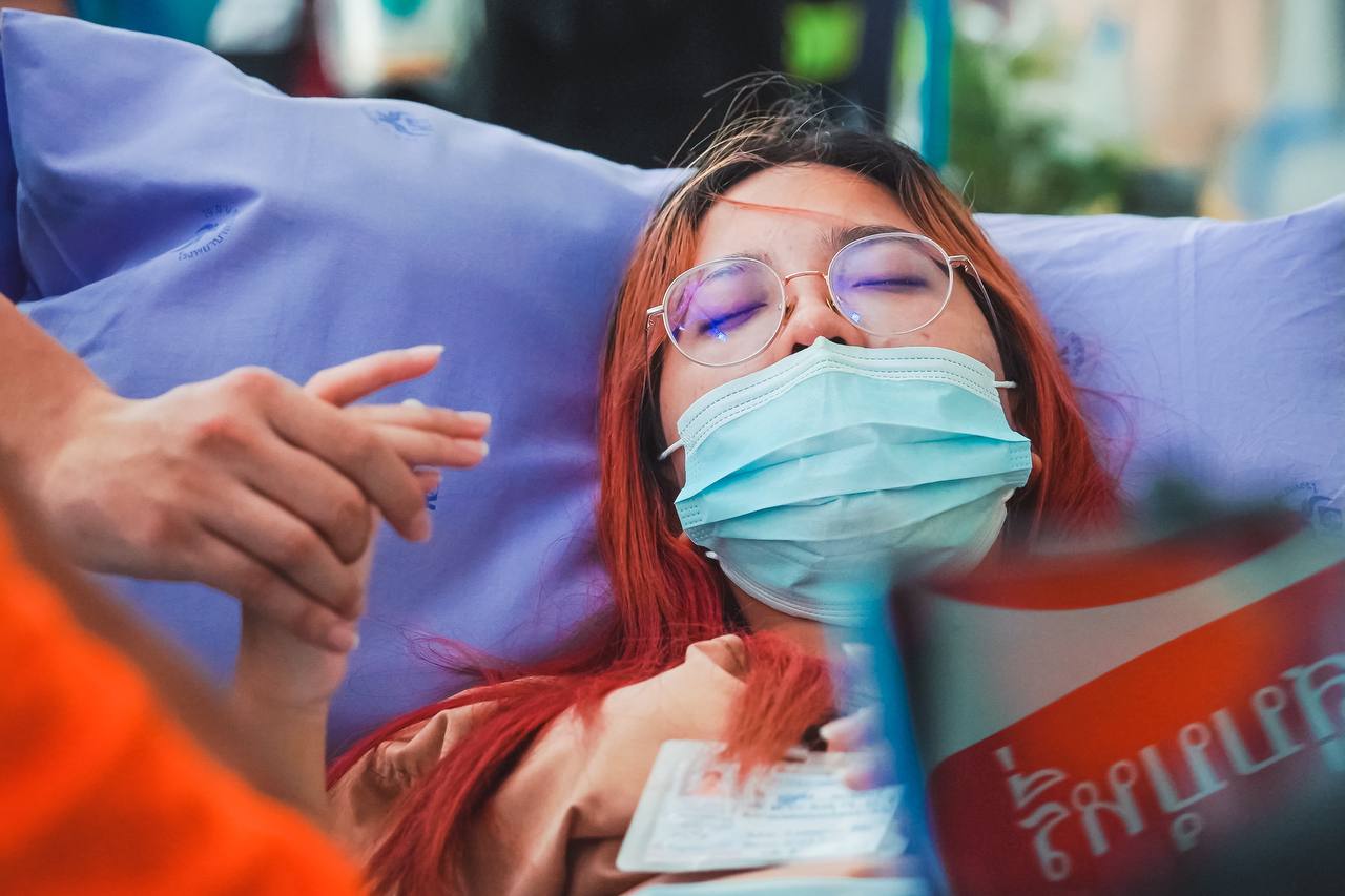 Das Bild zeigt eine Frau in einem Krankenbett mit geschlossenen Augen, jemand hält ihre Hand