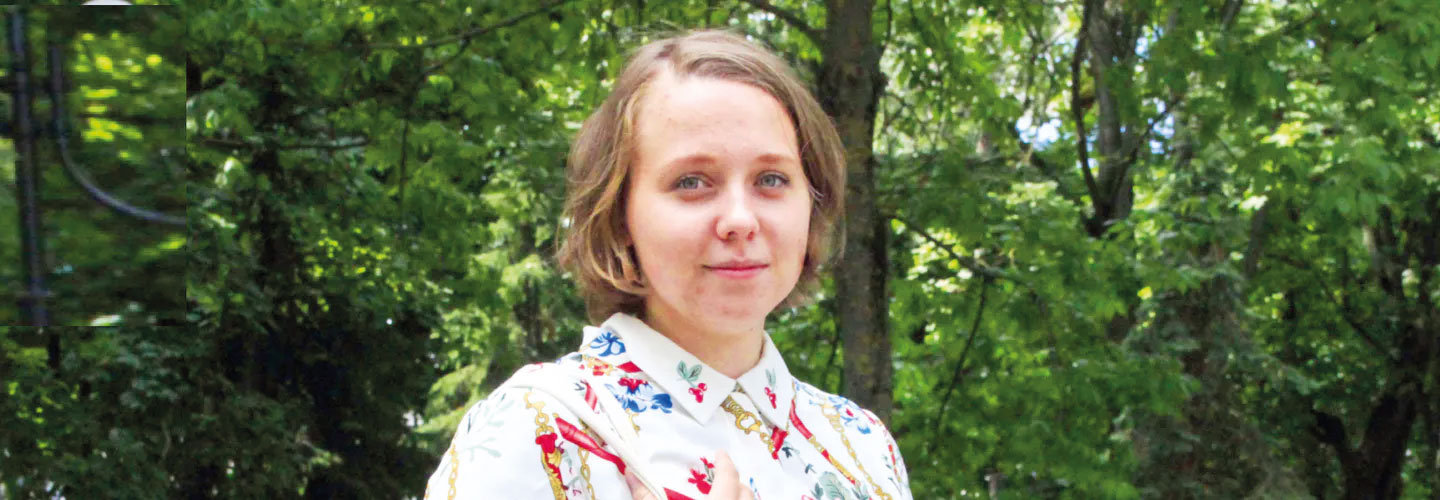 Eine junge Frau mit Haar, dass bis über ihre Ohren reicht, trägt eine Bluse, lächelt, steht vor einer Reihe von Bäumen und Büschen.