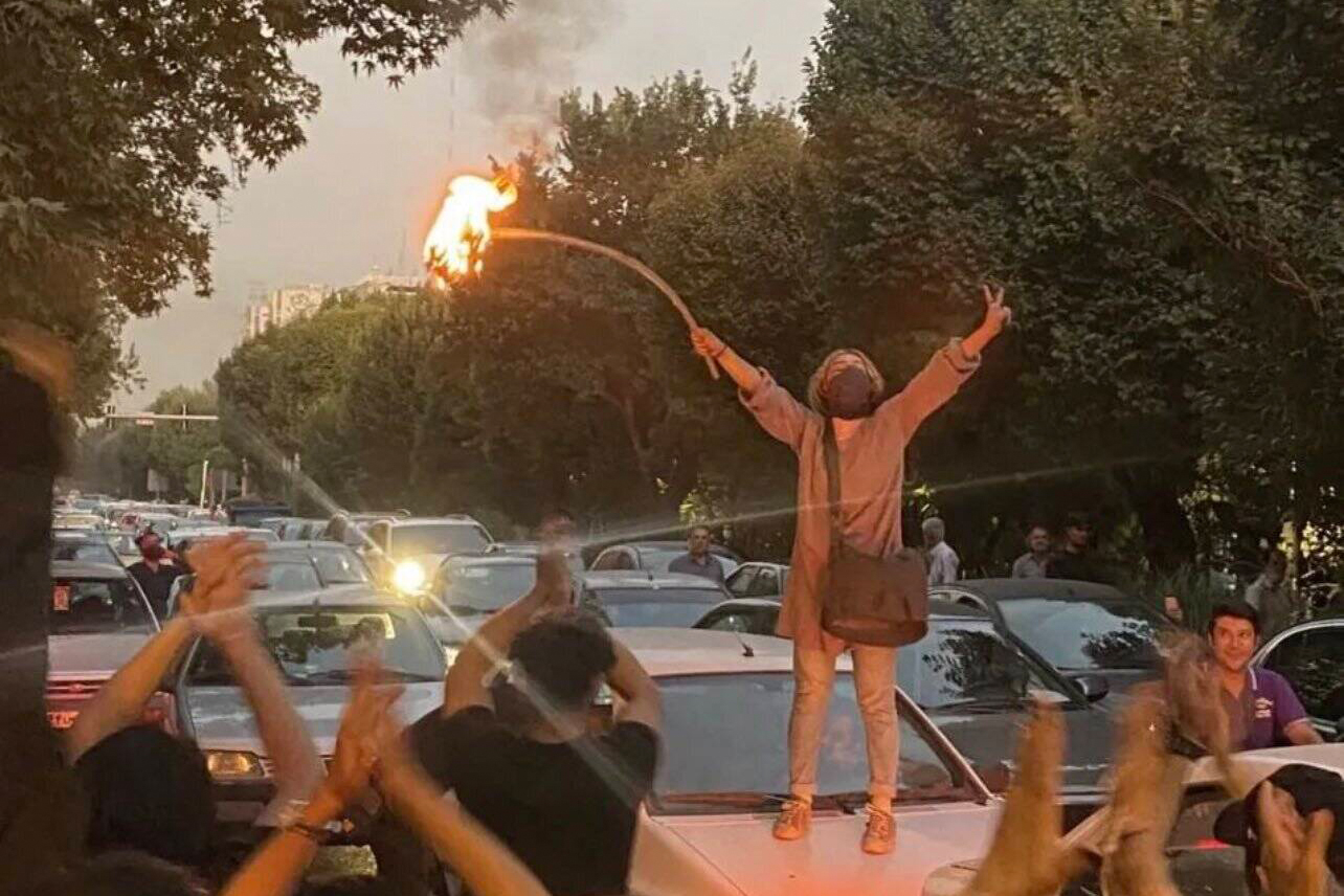 Das Bild zeigt das Porträtbild einer Frau, die mit einem brennenden Gegenstand auf einem Auto steht und die Hände in die Höhe hält.