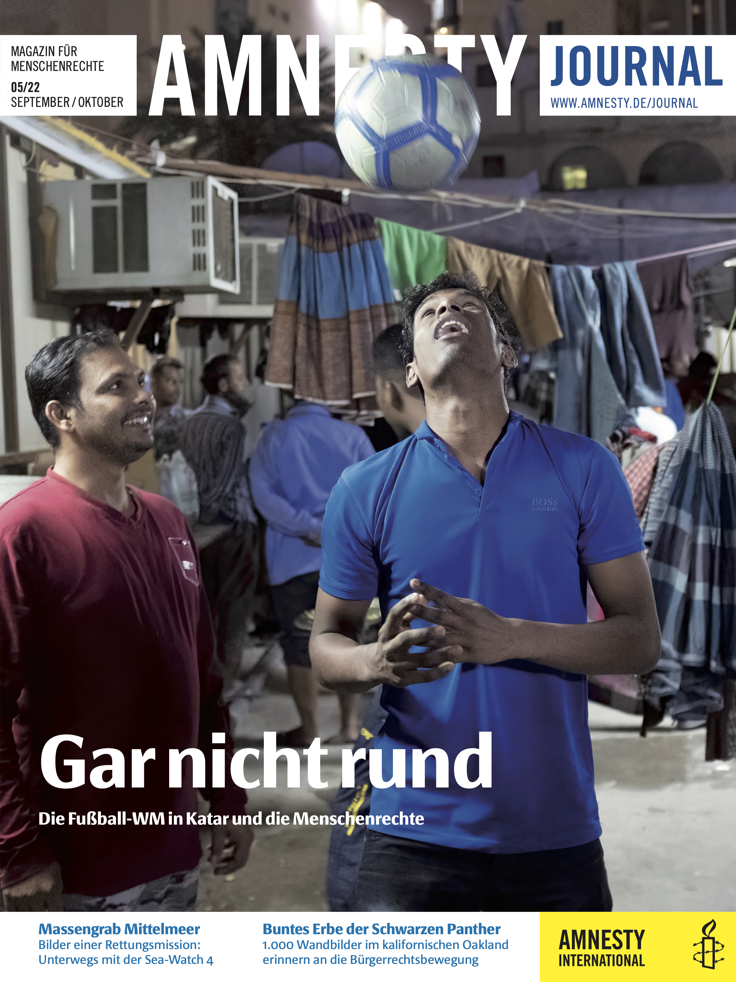 Zwei Männer stehen nebeneinander, der rechte von ihnen köpft einen Fußball in die Höhe, der andere beobachtet ihn dabei und lächelt; im Hintergrund sind große Stofftücher auf Wäscheleinen aufgehängt und Männer stehen beinander.