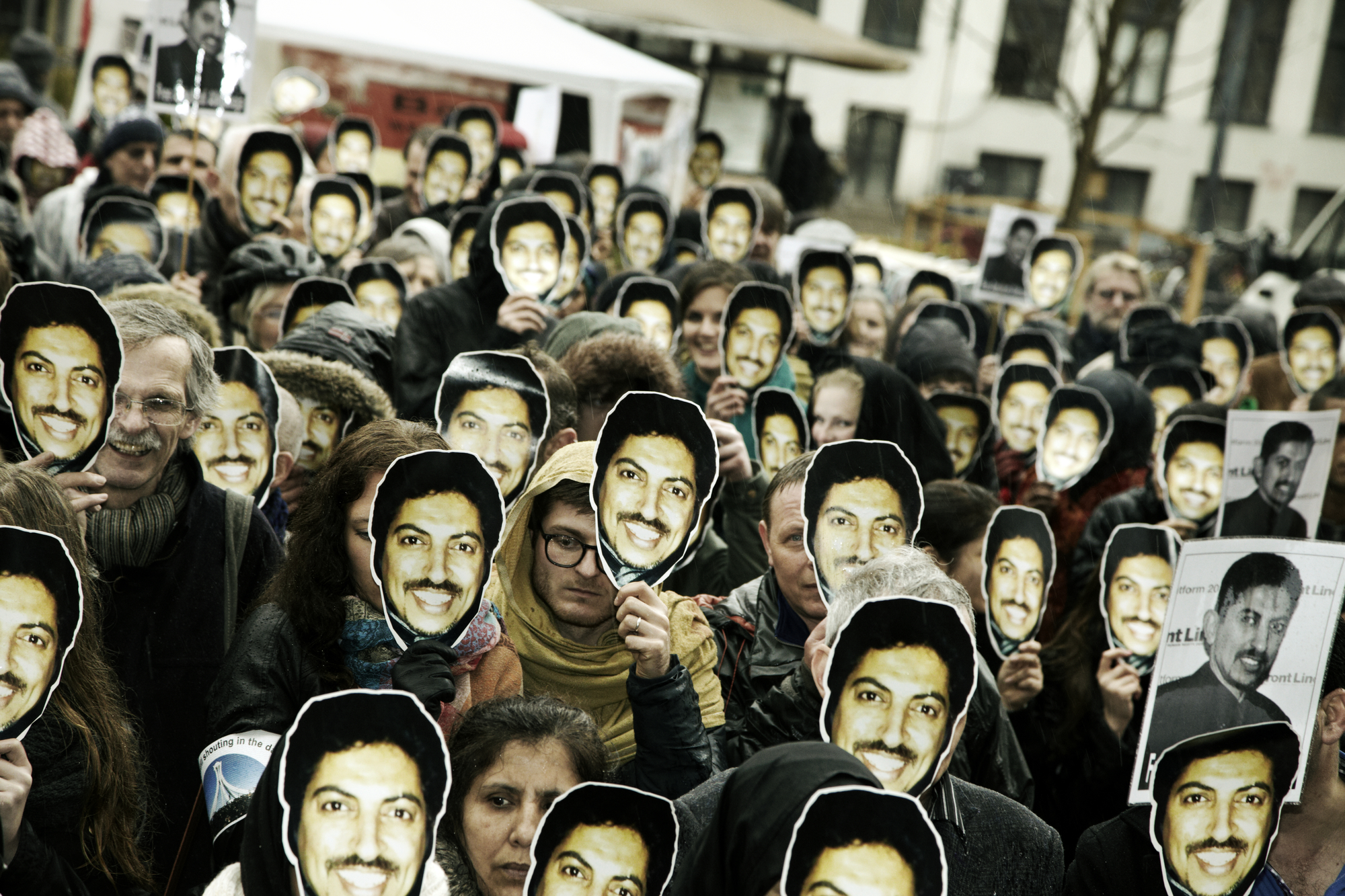 Dutzende Menschen stehen auf einem Platz und halten sich vor ihre Gesicher Masken mit dem Portätfoto des lächelenden Abdulhadi Al-Khawaja.