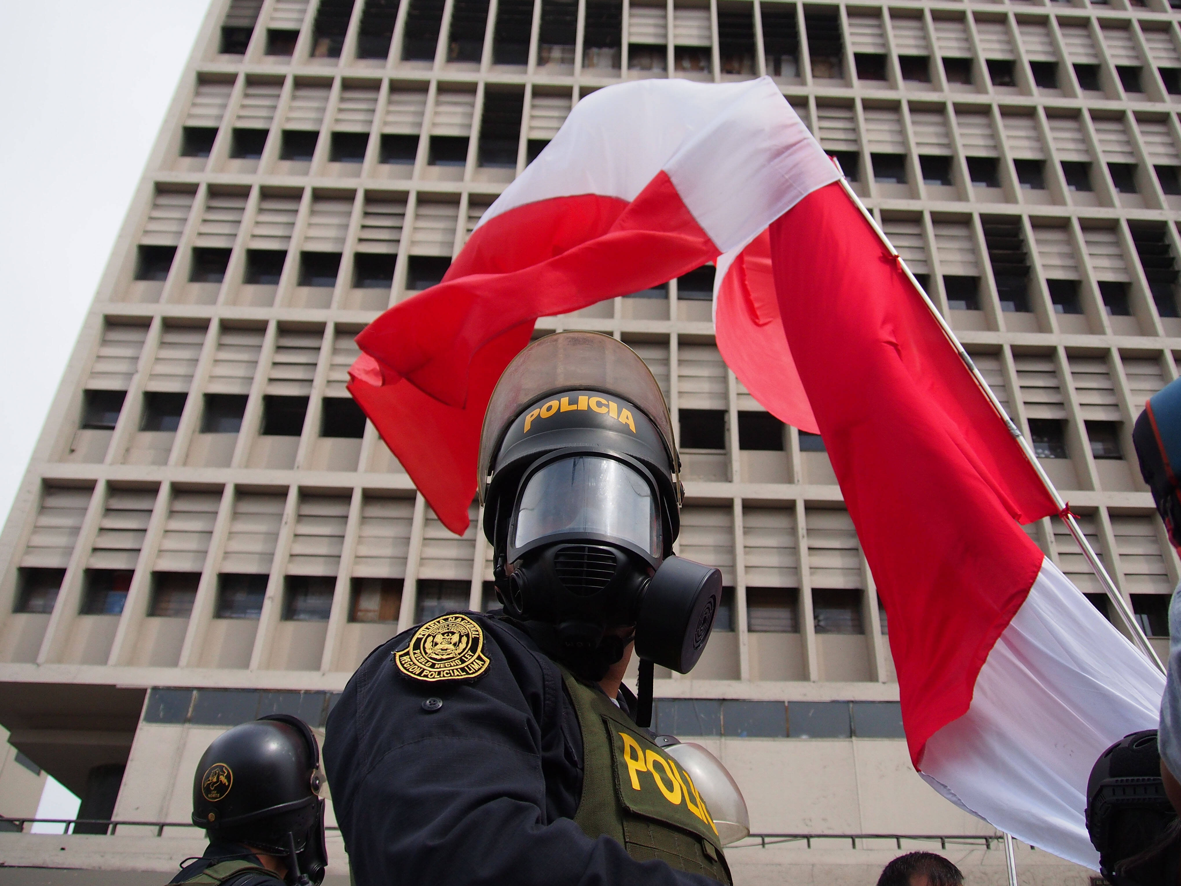 Da Bild zeigt im Vordergrund einen Polizisten mit ABC-Schutzmaske, dahinter die peruanische Flagge
