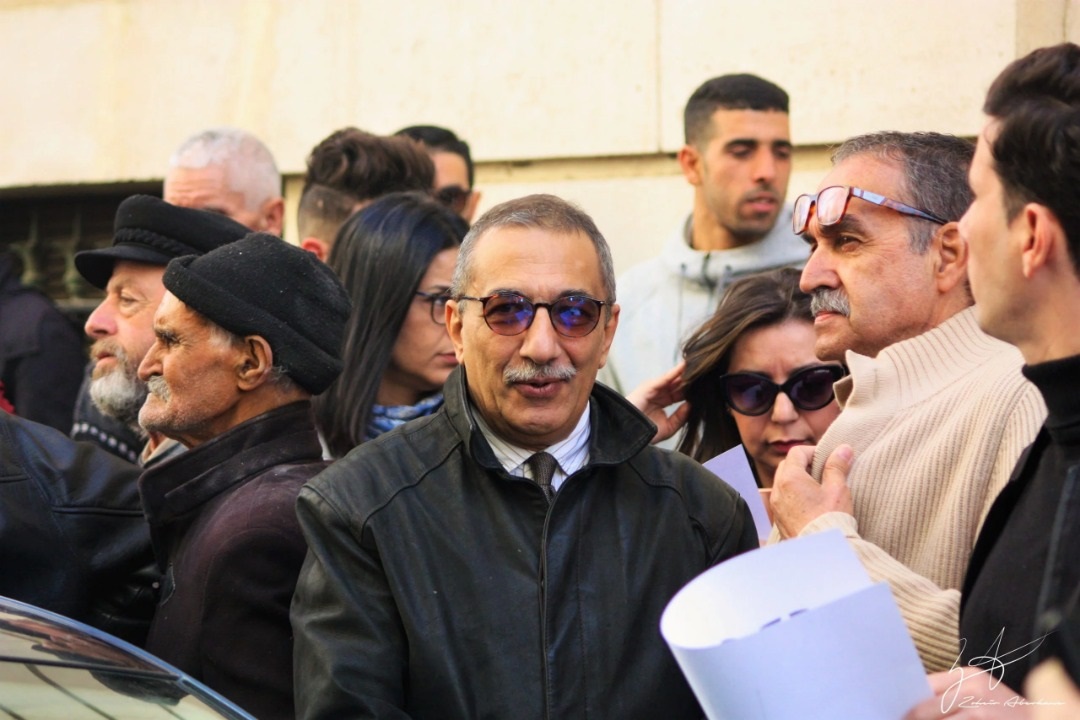 Das Foto zeigt Ihsane El Kadi, der einen Anzug mit Krawatte trägt und eine Sonnenbrille. Er steht umgeben von mehreren Personen vor einem Gebäude und blickt in die Kamera.