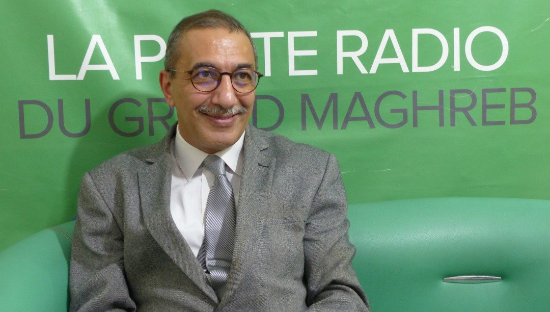 Das Foto zeigt Ihsane El Kadi lächelnd im Anzug mit Krawatte vor einem Banner sitzen, auf dem unter anderem die Wörter "Radio" und "Maghreb" stehen.
