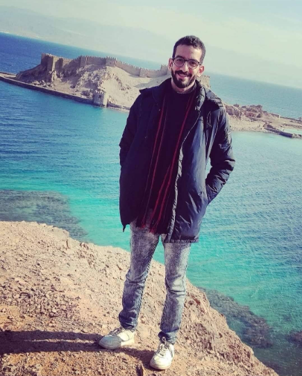 Das Bild zeigt das Porträtfoto eines Mannes auf einem Felsvorsprung, im Hintergrund das Meer, er lächelt in die Kamera