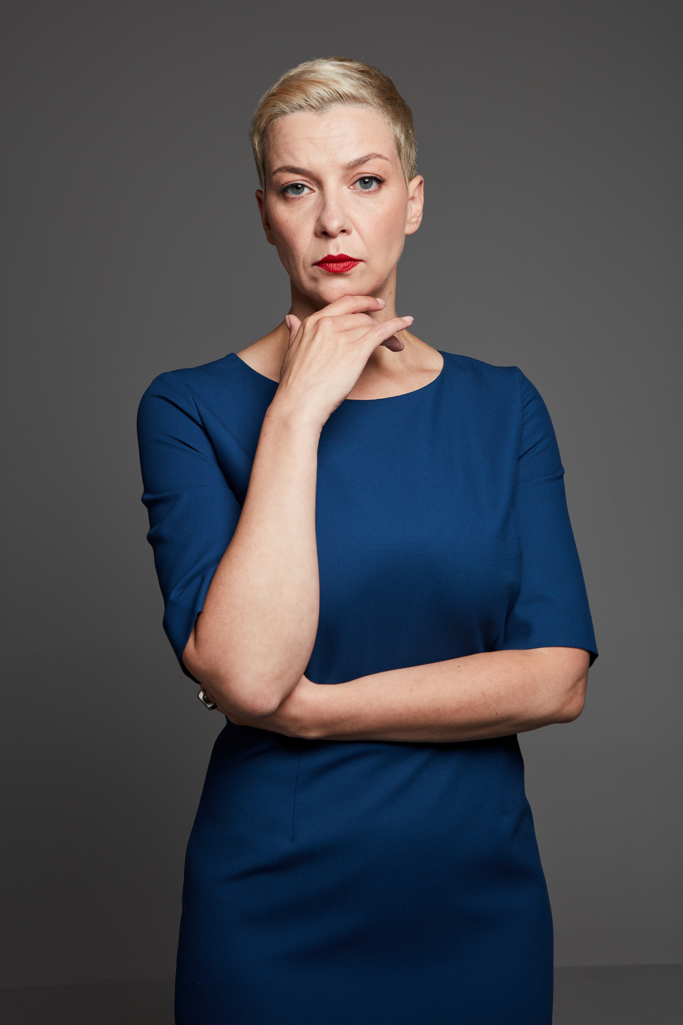Porträtaufnahme von Maryia Kalesnikava, die ein Kleid trägt und kurze blonde Haare und ernst in die Kamera schaut, während sie ihre recht Hand unter ihr Kinn hält.