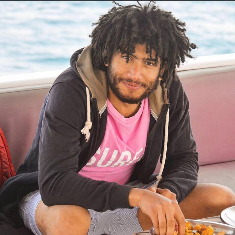 Foto von Badr Mohamed, der an einem Gewässer in die Kamera lächelnd auf einer Bank sitzt und mit der rechten Hand nach Essen greift.