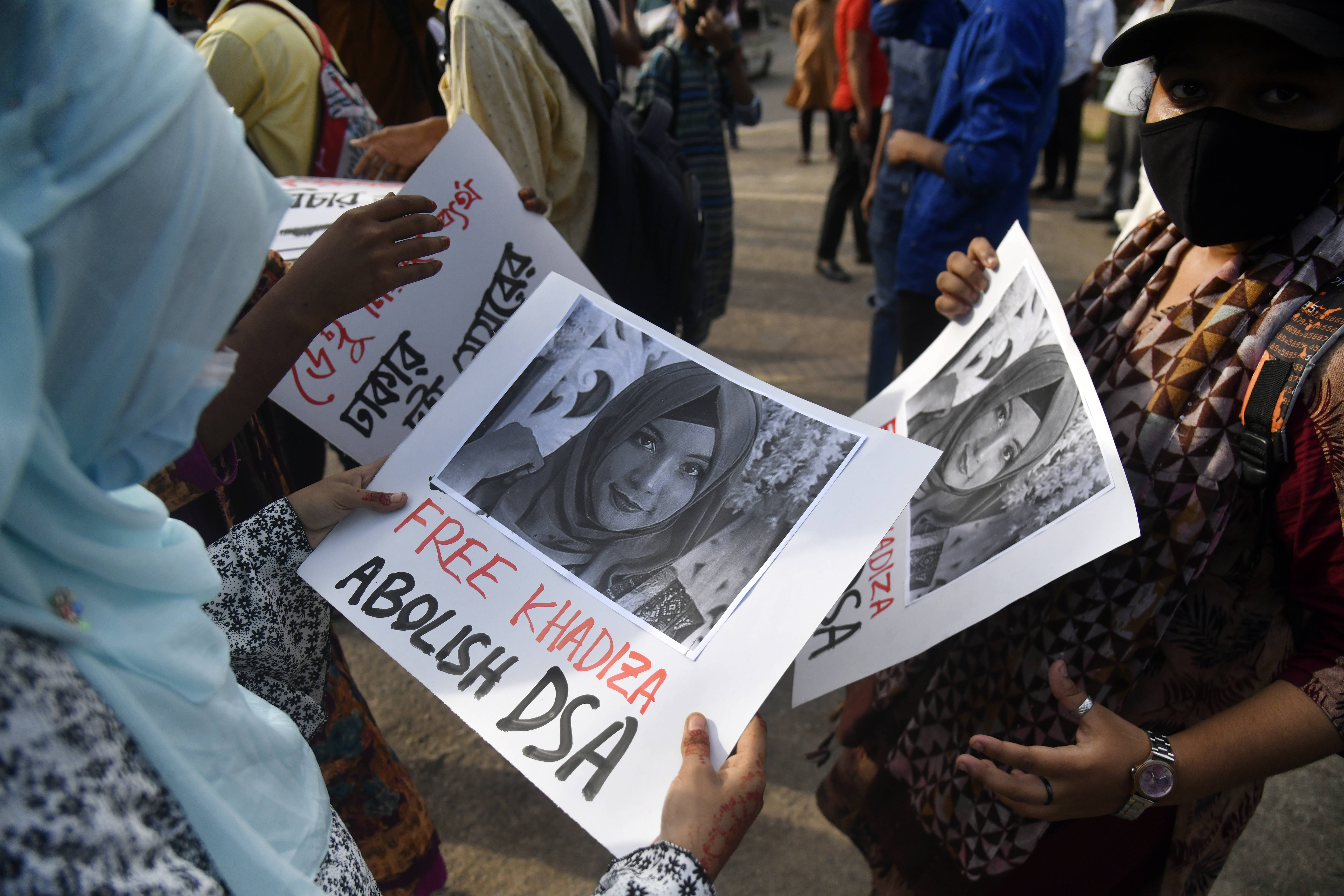 Eine Frau steht in einer Menschenmenge. Sie hält ein Plakat in ihren Händen und betrachtet ist. Auf dem Plakat ist ein Porträtfoto von Khadijatul abgedruckt, auf dem sie ein Kopftuch trägt. Unter dem Foto steht unter anderem: "Free Khadiza".