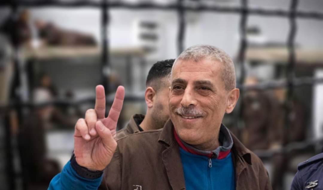 Ein älterer Mann mit kurzen grauen Haaren lächelt und formt mit Zeige- und Mittelfinger ein "V", es ist Walid Daqqah. 