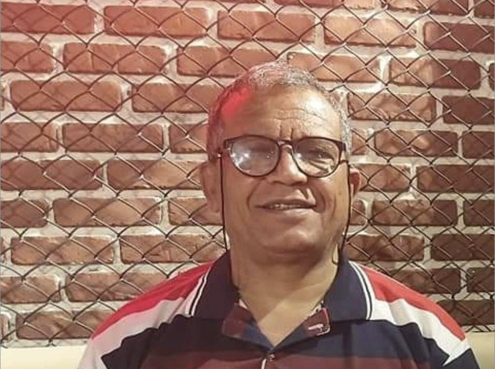 Portraitaufnahme von Abdul-Baqi Saeed, der vor einer Ziegelmauer steht und eine Brille und ein Polo-Hemd trägt und in die Kamera lächelt.