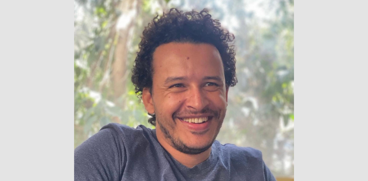 Ein junger Mann mit kurzen, lockigen, schwarzen Haaren lächelt. Es ist Mahmoud Hussein. Im Hintergrund Bäume. 