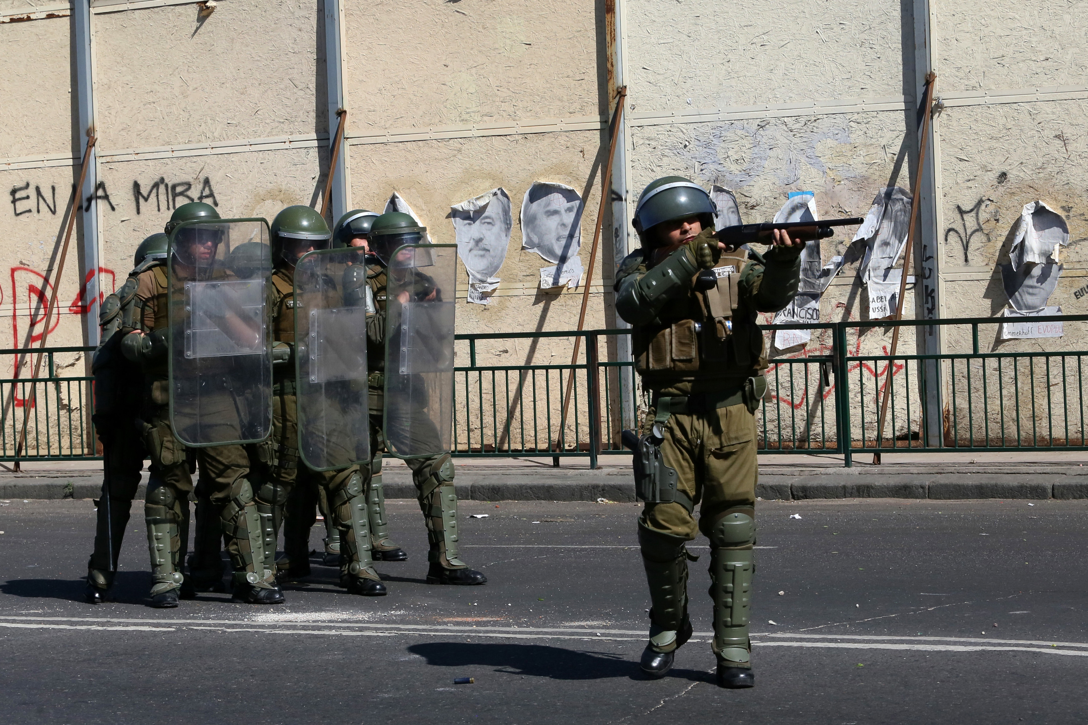 Das Foto zeigt eine Gruppe von sechs Polizisten mit Helmen und Schutzschildern. Vor ihnen steht ein weiterer Polizist mit Helm, der mit einem Gewehr anlegt und zielt.