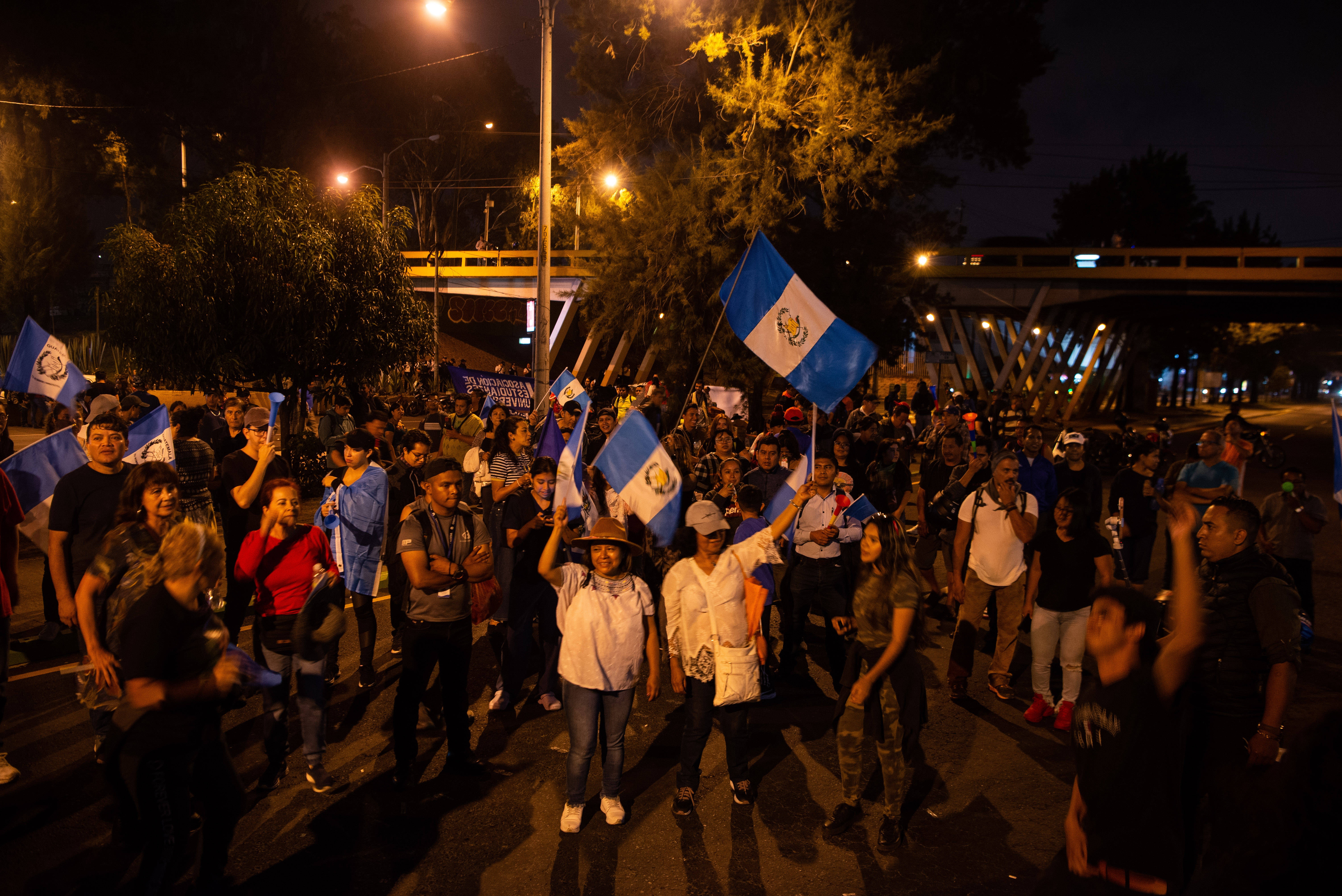 Das Foto zeigt eine große Gruppe Demonstrierender bei Nacht auf einer Straße. Einige von ihnen schwenken die Flagge Guatemalas.