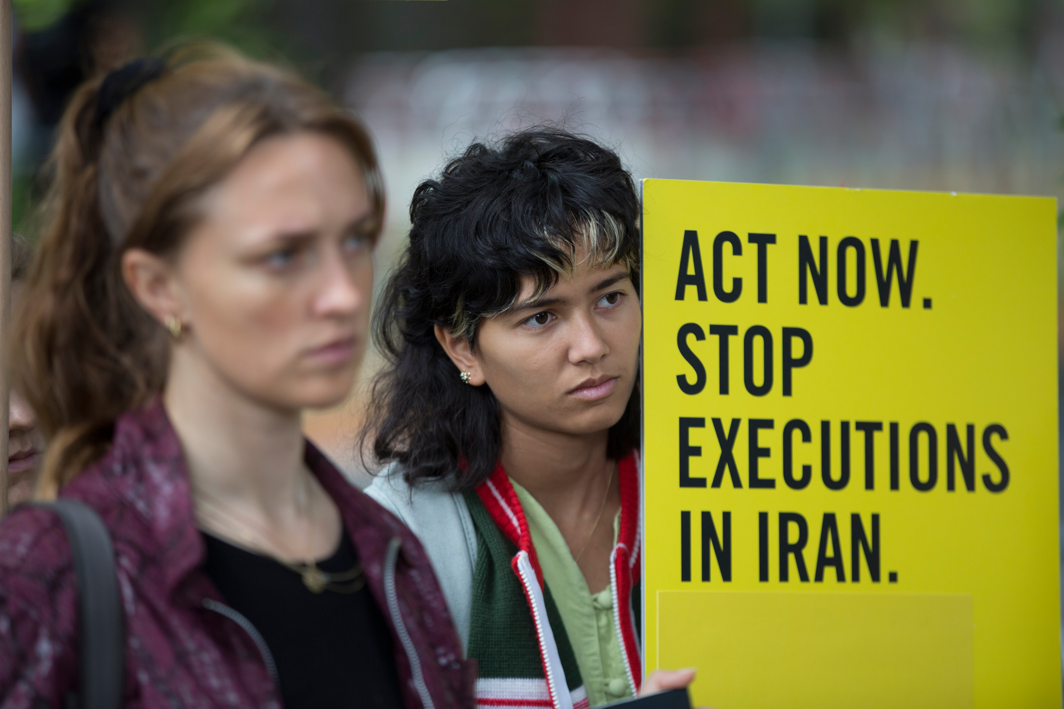 Das Bild zeigt zwei Frauen, die beide ernst schauen und von denen eine, ein gelbes Schild mit der schwarzen Aufschrift "Act now. Stop executions in Iran." hält.