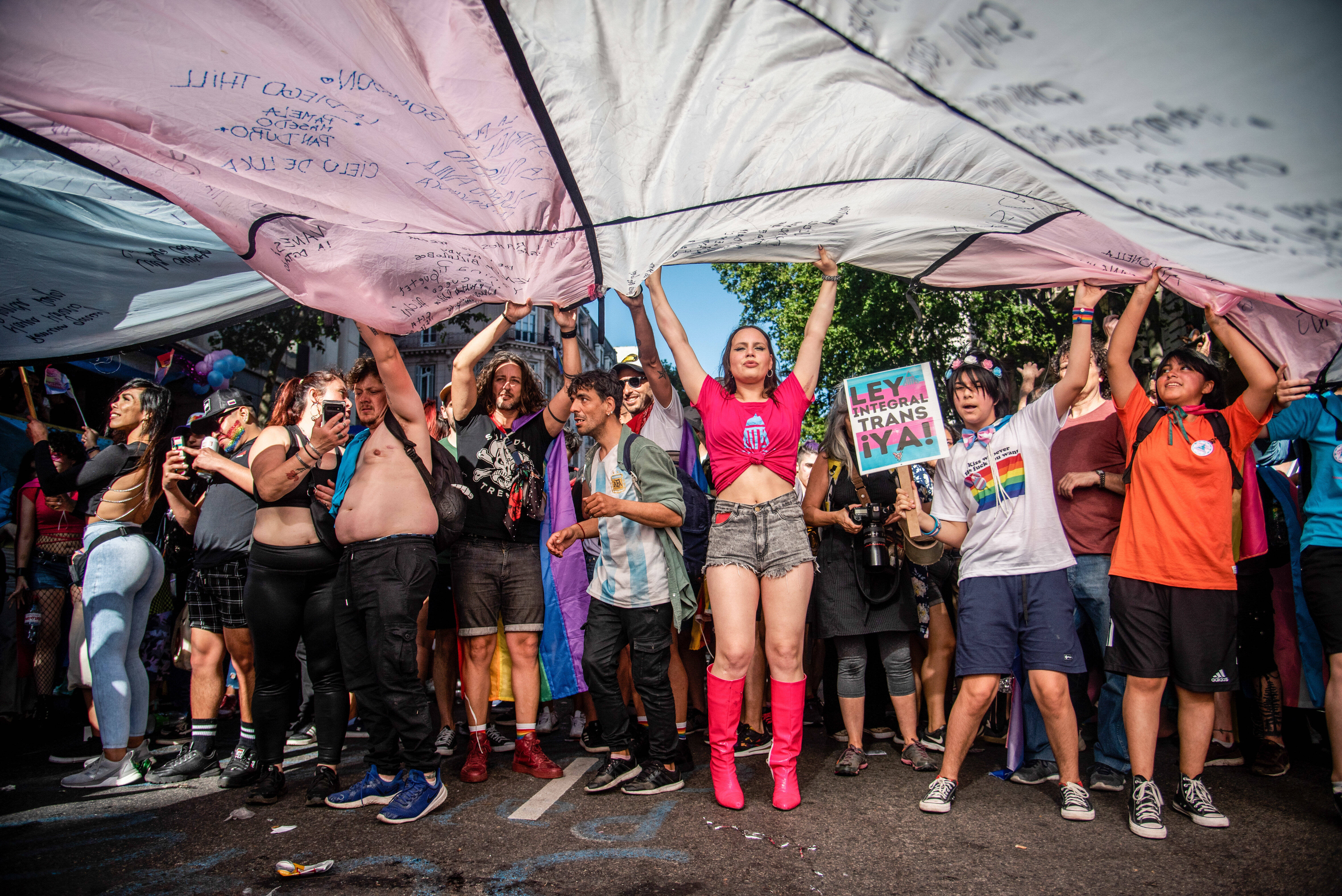 Mehrere Personen stehen auf einer Straße nebeneinander und breiten über ihren Köpfen eine große Transgender-Pride-Fahne aus.