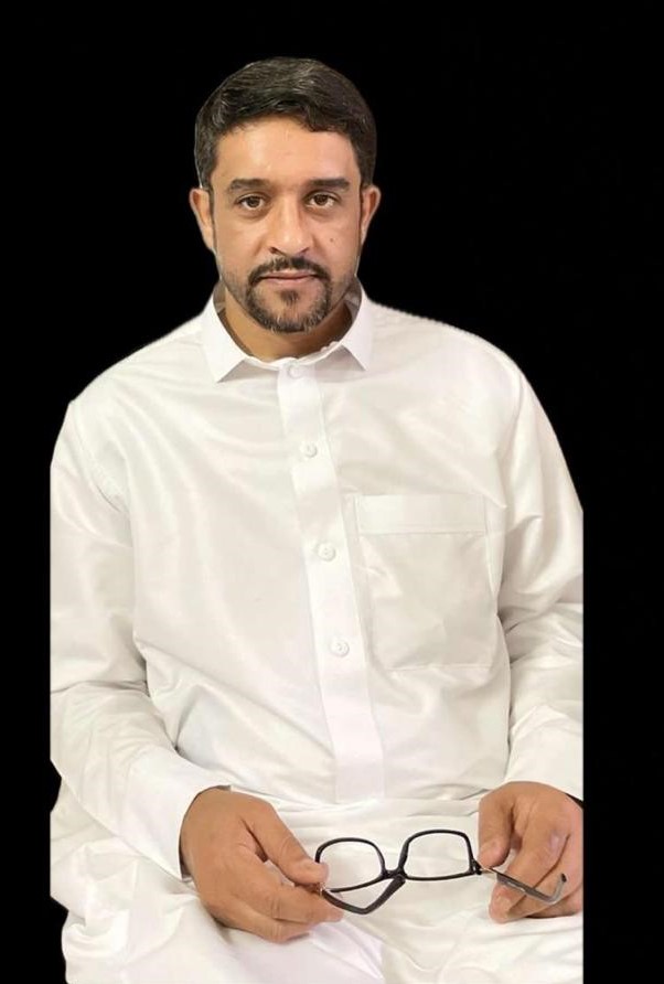 Das Foto zeigt Asaad bin Nasser al-Ghamdi, der ein langes Gewand trägt und seine Brille in der rechten Hand auf seinem Schoß hält und in die Kamera lächelt.